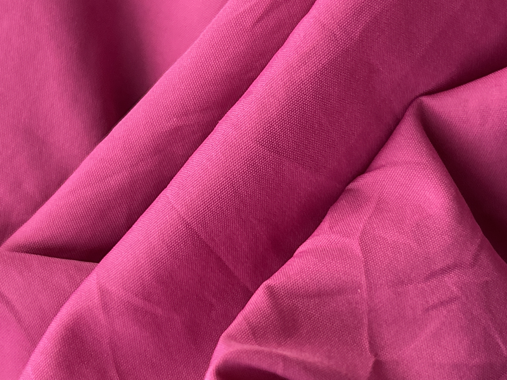 This is a close-up, overhead shot of a vibrant fuchsia pink fabric. The fabric is draped and folded, creating a textured surface with shadows and highlights that emphasize the folds. The weave of the fabric is visible, appearing to be a tightly woven material. The lighting suggests an indoor setting, and there are no people or other objects present. The image does not contain any information related to the city of Phuket City Municipality, Thailand.