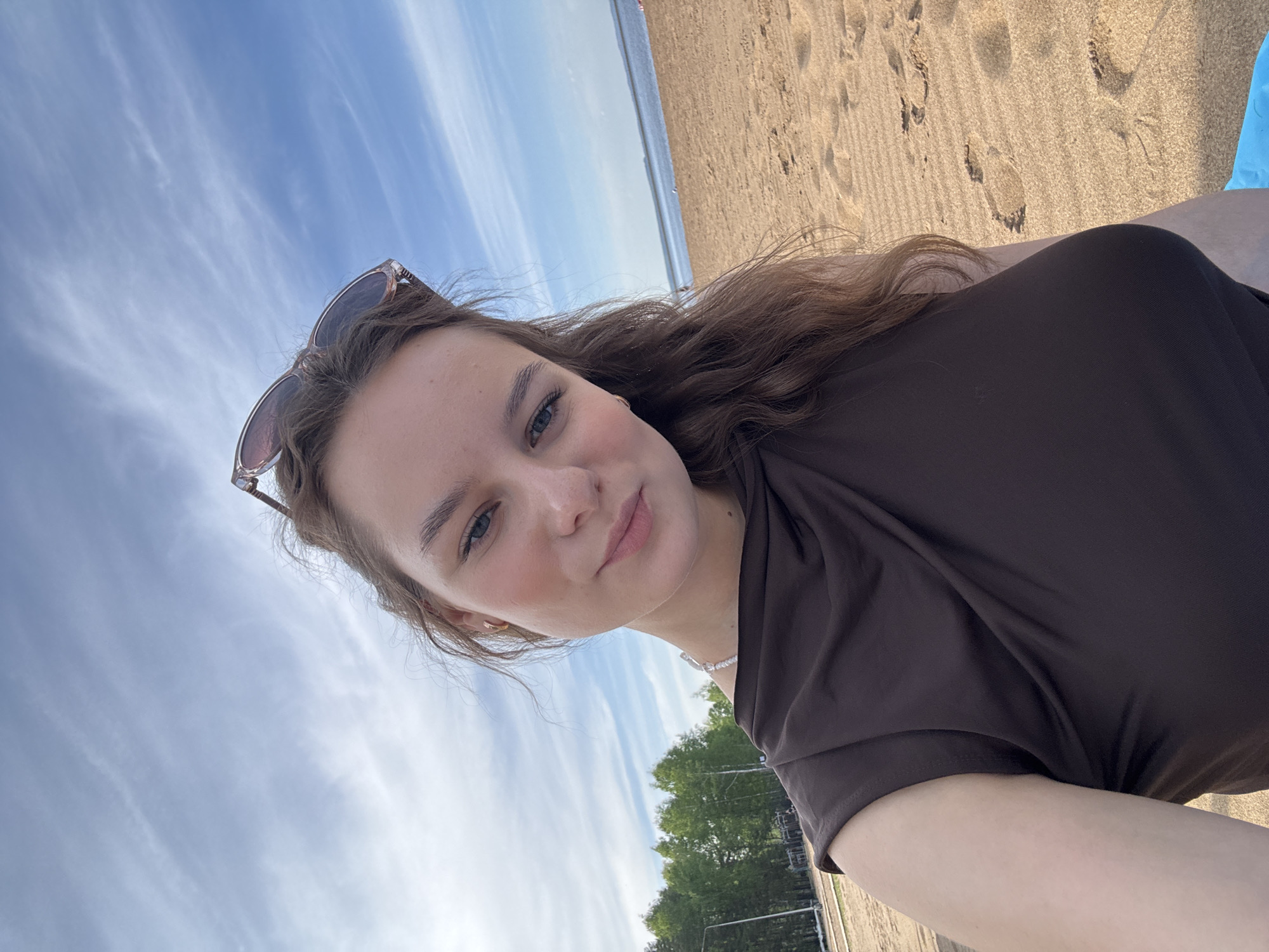 A young woman with light skin and brown hair is taking a selfie on what appears to be a beach. She is wearing a brown top and has sunglasses perched on her head. In the background, there is a sandy beach with footprints, a body of water, and a blue and white cloudy sky. Green trees and a portion of what seems to be a fence are visible in the lower left. Based on the prompt, the location is likely Chaikovsky, Russia.