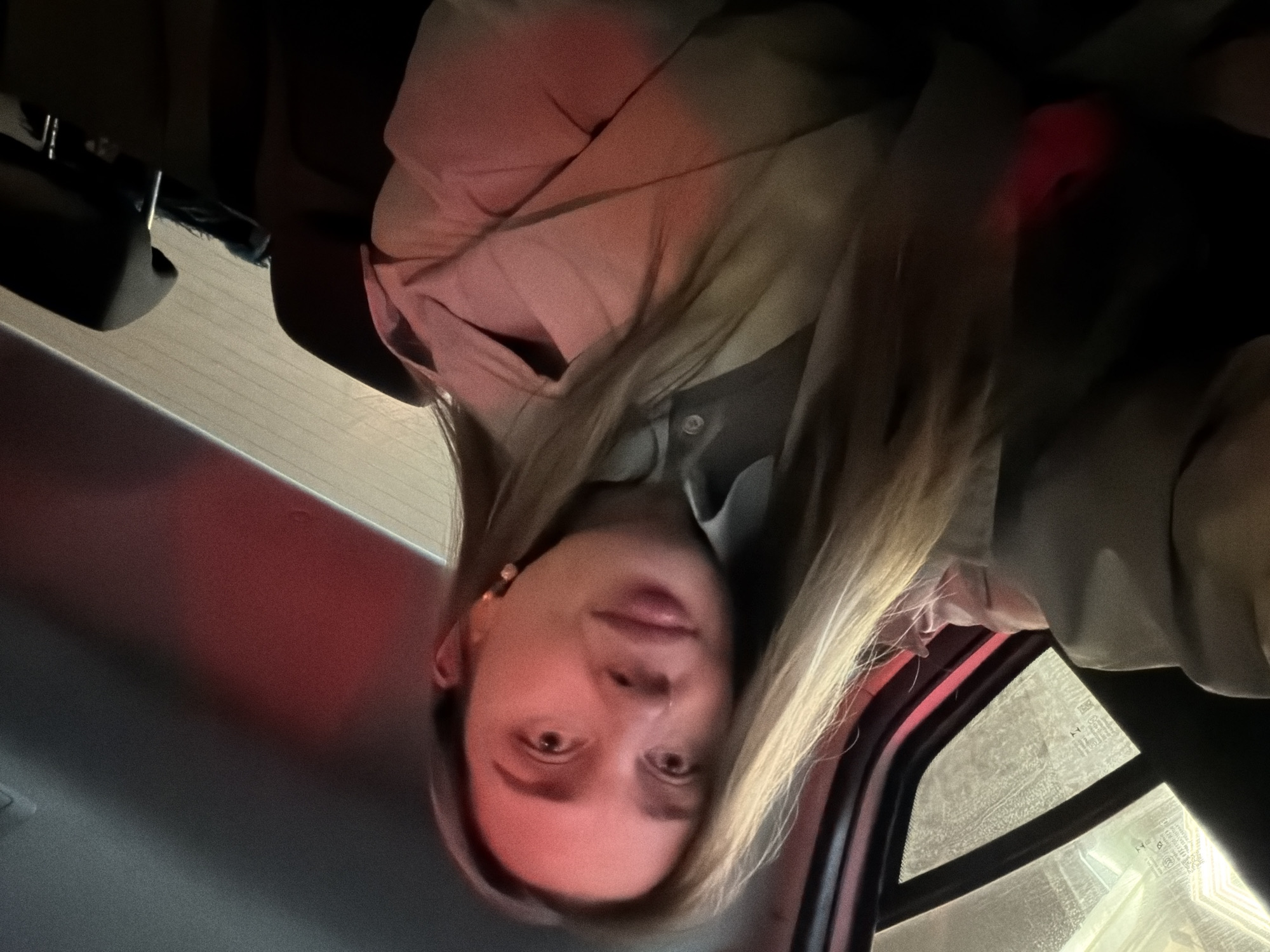 The image depicts a woman inside a car. The car appears to be at a standstill. The woman has blonde hair and is wearing a tan jacket. The environment is dark, and there is red light present, possibly from the car's tail lights or another vehicle nearby. Based on the metadata, the location is Moscow, Russia. There are no visible interactions or actions taking place.
