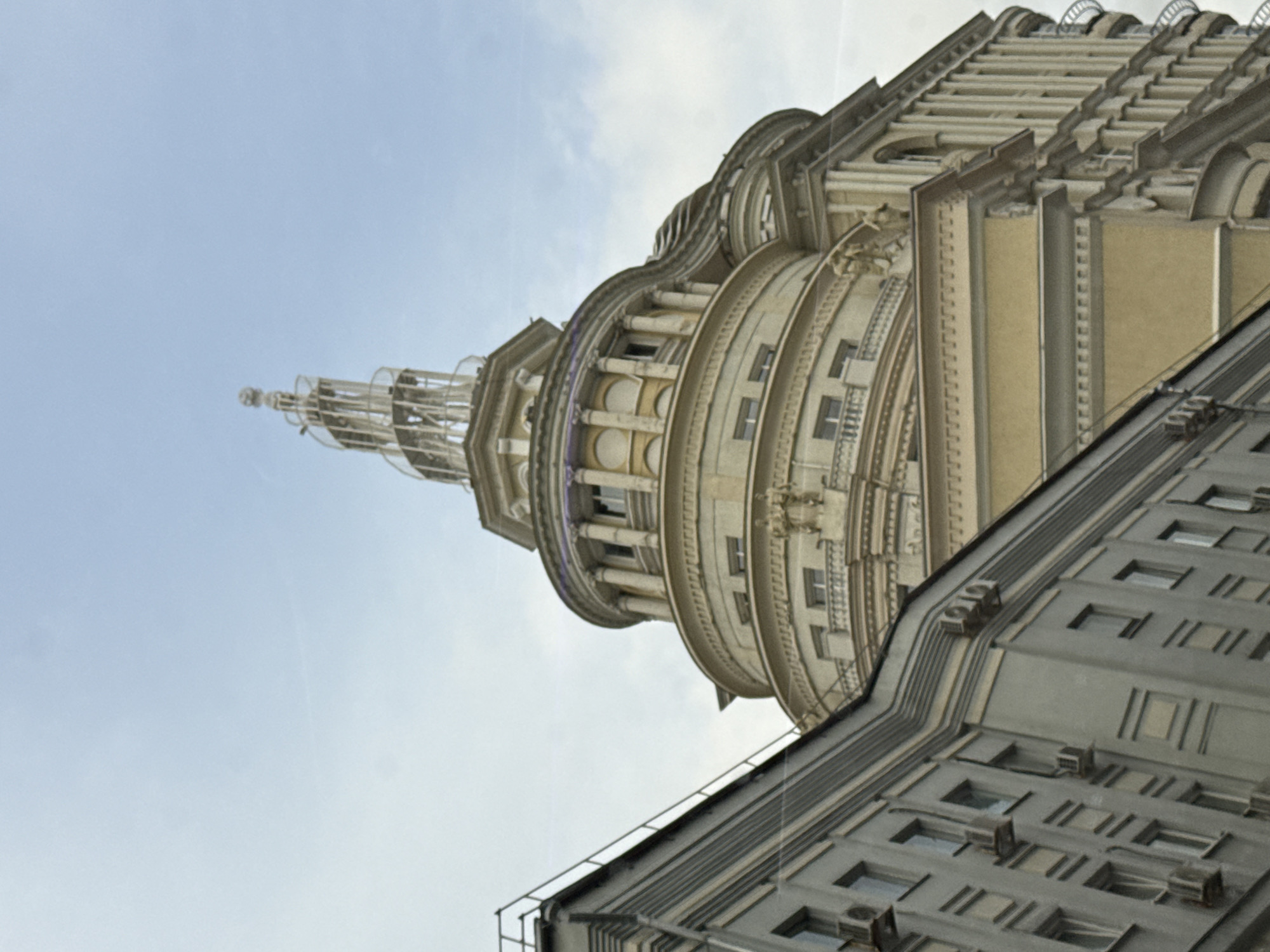 The image shows a building in Moscow, Russia, viewed from a low angle. The building has a multi-tiered, ornate tower with a spire at the top. The tower is constructed in a circular or rounded form with columns and decorative elements. The facade is light-colored, likely stone or stucco. Attached to the tower is a less ornate, rectangular section of the building with evenly spaced windows and air conditioning units on the roof. The sky is overcast with a pale blue color.