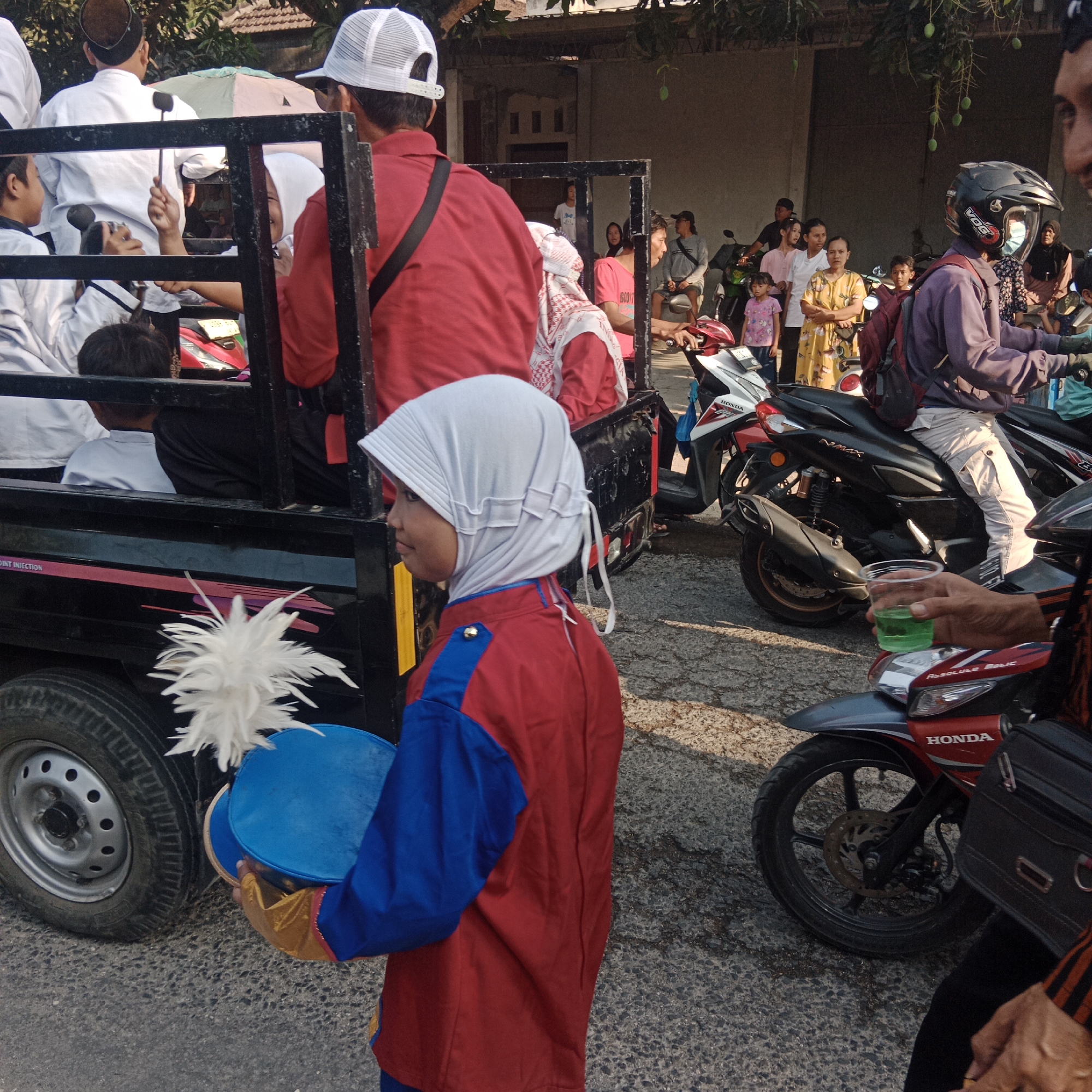 The image captures a street scene in Tegalrejo, Indonesia, likely during a parade or cultural event. In the foreground, a young person wearing a white hijab and a red and blue uniform is holding a blue drum and a white feathered baton, suggesting participation in a marching band or performance group. Behind them, a black pickup truck bed is visible, containing several individuals dressed in white attire and red shirts. Some are wearing white caps. The truck is parked on an asphalt road.
Further in the background, a crowd of people is gathered on the side of the road. Many are standing around parked motorcycles. Several individuals are dressed in casual clothing, with some women wearing headscarves. A few people are seated on motorcycles, and one person is holding a clear plastic cup with green liquid. The environment appears to be a street lined with buildings, some with visible windows and doorways, and trees with hanging fruit, possibly mangoes, suggesting a tropical or subtropical climate. The overall lighting indicates it is daytime, with sunlight casting shadows. The presence of multiple motorcycles and the gathering of people suggest a community event or a public celebration.