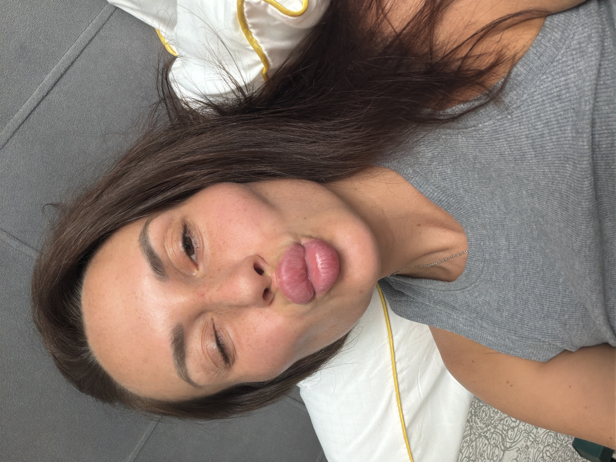 The image shows a woman with dark brown hair and fair skin, lying down and making a kissing face. She is wearing a grey tank top and a thin silver necklace. Her eyes are closed and her lips are pursed. She is lying on a grey surface, possibly a couch or bed, with a white pillow with a yellow trim behind her head. The setting appears to be indoors, and there are no immediate visual cues to suggest a specific location within Moscow, Russia.