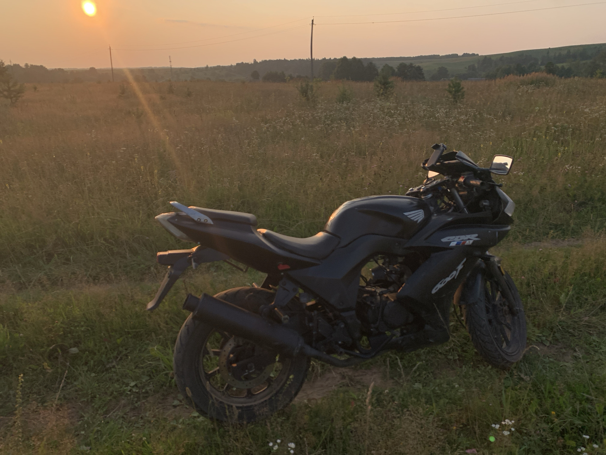 A black motorcycle is parked on a dirt path in a field of tall grass and wildflowers, under a warm, orange sunset sky. The motorcycle is angled to the right, with its front wheel turned slightly. The field stretches out behind the motorcycle, with a line of trees and hills in the distance. A power line crosses the sky. The location is identified as the city of Panteleevka, Russia.