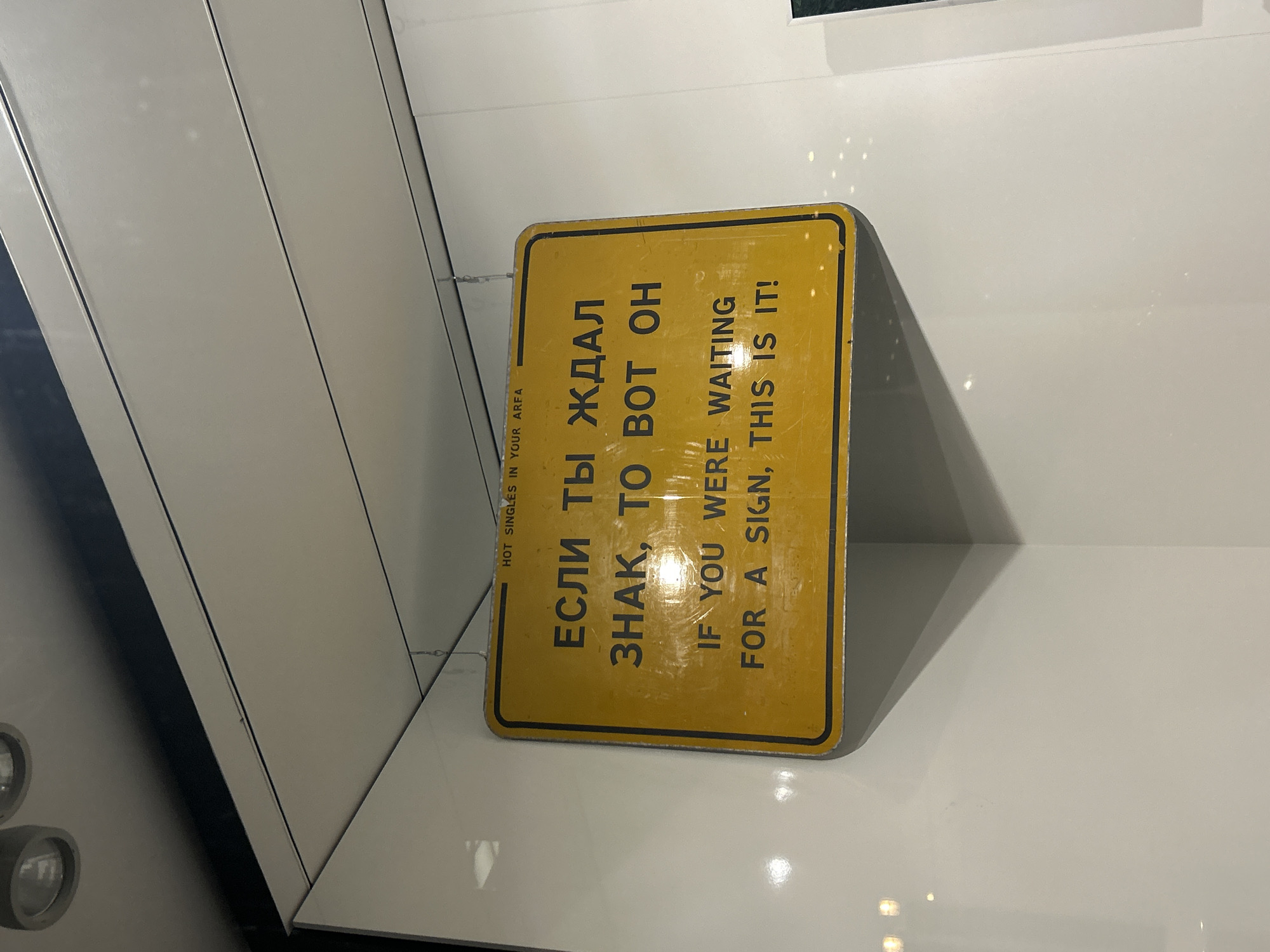 The image shows a yellow sign with black text in both Russian and English that reads "ЕСЛИ ТЫ ЖДАЛ ЗНАК, ТО ВОТ ОН IF YOU WERE WAITING FOR A SIGN, THIS IS IT!". The sign is slightly angled and appears to be inside a museum or exhibit in Berlin, Germany. The background is white, and there's a black ledge or panel to the left.