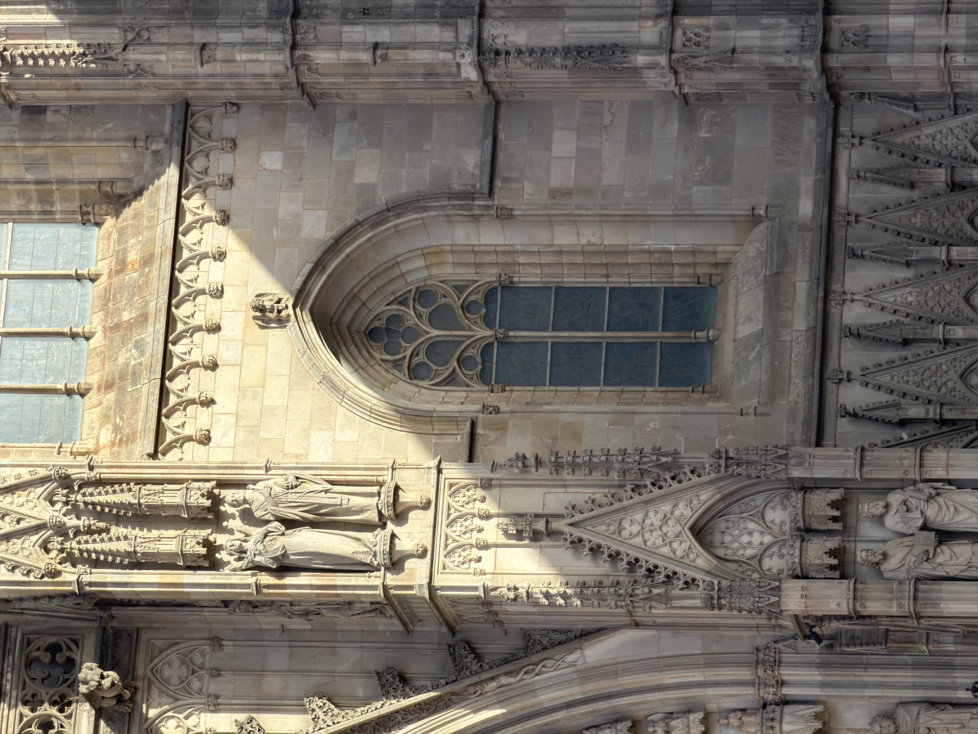 The image shows a detail of the facade of the Barcelona Cathedral, located in the Gothic Quarter of Barcelona, Spain. The architecture is Gothic, characterized by ornate stone carvings, pointed arches, and intricate tracery.
In the center of the image, a large, pointed arch window with a complex tracery design is visible. The window is divided into multiple panes, and a bluish-green tint suggests it might be stained glass, though the detail is not sharp enough to confirm definitively. The window is set into a wall of light-colored, roughly cut stone blocks.
To the left of the window, a section of the facade features two large stone statues of figures in draped robes. They appear to be saints or biblical figures, with solemn expressions. Above them, more elaborate stone carvings are present, including smaller figures and decorative architectural elements. To the left of these statues, a darker arched doorway or niche is visible, with more statuary within, partially obscured by shadow.
To the right of the central window, the facade continues with vertical stone elements that are heavily decorated with Gothic ornamentation, including small pinnacles and tracery. This section is cast in shadow, revealing the depth and texture of the carvings.
The overall scene is bathed in sunlight, creating strong contrasts between light and shadow, which highlights the texture and detail of the stonework. The image focuses on a specific section of the cathedral's exterior, emphasizing its architectural grandeur and sculptural richness.