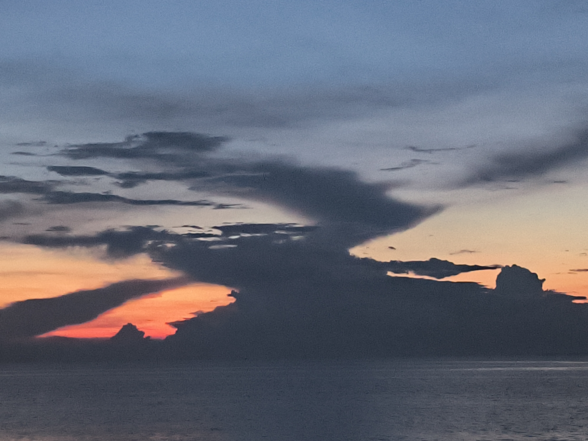 The image captures a seascape under a dynamic sky. The scene is likely during either sunrise or sunset, with an orange glow visible beneath dark, layered clouds that stretch across the horizon. The clouds have varied shapes and densities, some appearing wispy and translucent while others are thick and opaque. The water is calm, reflecting the colors of the sky.
There is no specific information in the image to confirm the location as Lhokseumawe, Indonesia. Without additional context or details, it's impossible to verify that this image was taken in that particular city. The scene depicts a natural environment with no people or identifiable objects to indicate a specific location.
