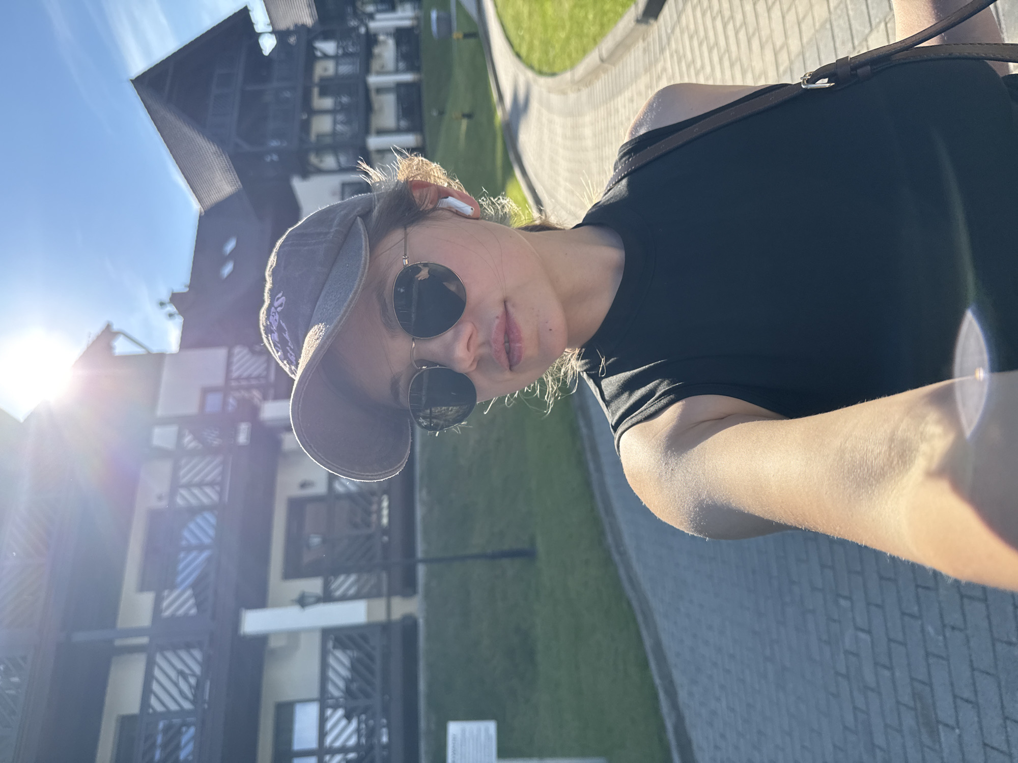 A young woman wearing a black sleeveless top, a grey baseball cap, and sunglasses is taking a selfie outdoors. She has a white wireless earbud in her right ear and a brown strap of a bag visible on her shoulder. The background shows a paved walkway, a grassy area, and buildings with wooden balconies. The sun is shining brightly, creating a lens flare in the upper left corner. The architecture of the buildings suggests a location in a mountainous or resort area, consistent with Szklarska Poręba, Poland.