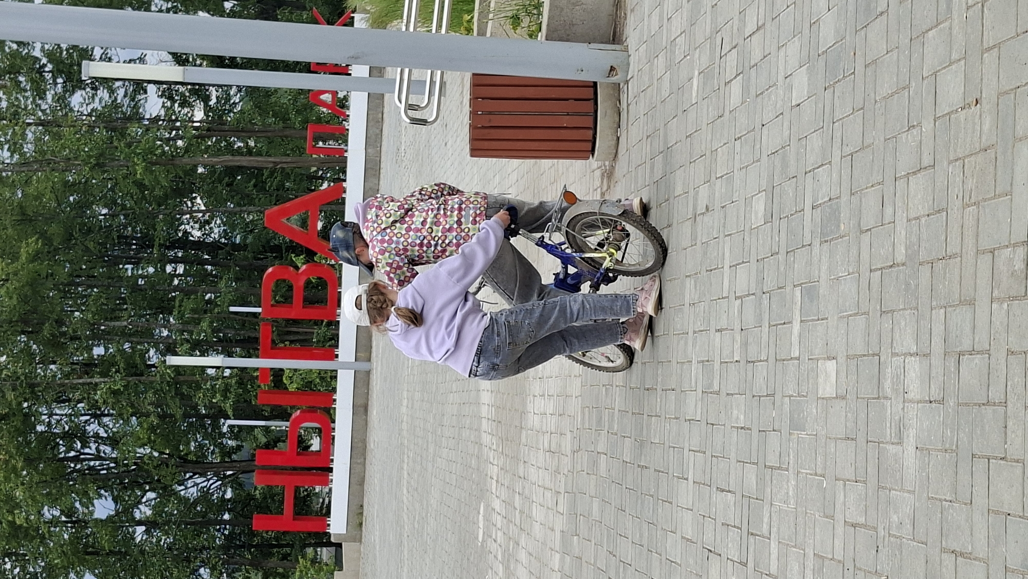Two children, a girl and a boy, are interacting with a blue bicycle on a paved walkway. The girl, wearing a light purple hooded sweatshirt and blue jeans, is on the left and appears to be helping the boy, who is on the right. The boy is wearing a colorful patterned hooded sweatshirt and a dark baseball cap. He is seated on the bicycle, and the girl has her hands on the handlebars and the back of the bike, suggesting she is either teaching him to ride or assisting him. They are positioned in front of a large red sign that spells out "НЫТВА" (Nytva) in bold, block letters, indicating this is likely a welcome sign or landmark in the city of Nytva, Russia. The background consists of a line of trees and a cloudy sky. To the right of the children, there is a wooden trash receptacle and a silver railing. The ground is paved with light gray rectangular bricks arranged in a repeating pattern.