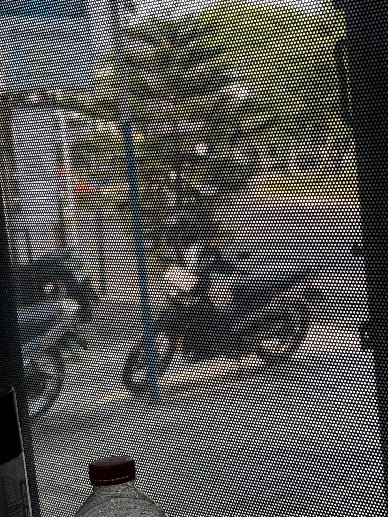 The image depicts a view through a perforated screen, likely a window film, looking outwards. In the foreground, the top of a clear plastic water bottle with a dark red cap is visible, along with a portion of a spray bottle labeled "800ml." The background shows a sunlit outdoor scene. Through the perforations, several motorbikes are discernible, suggesting a street or parking area. The environment appears to be a city, as indicated by the presence of vehicles and a general urban setting. The overall lighting suggests it is daytime, with bright sunlight filtering through the foliage and casting shadows. The location is identified as Kepolorejo, Indonesia. The scene is captured from a static viewpoint, looking through the screen.