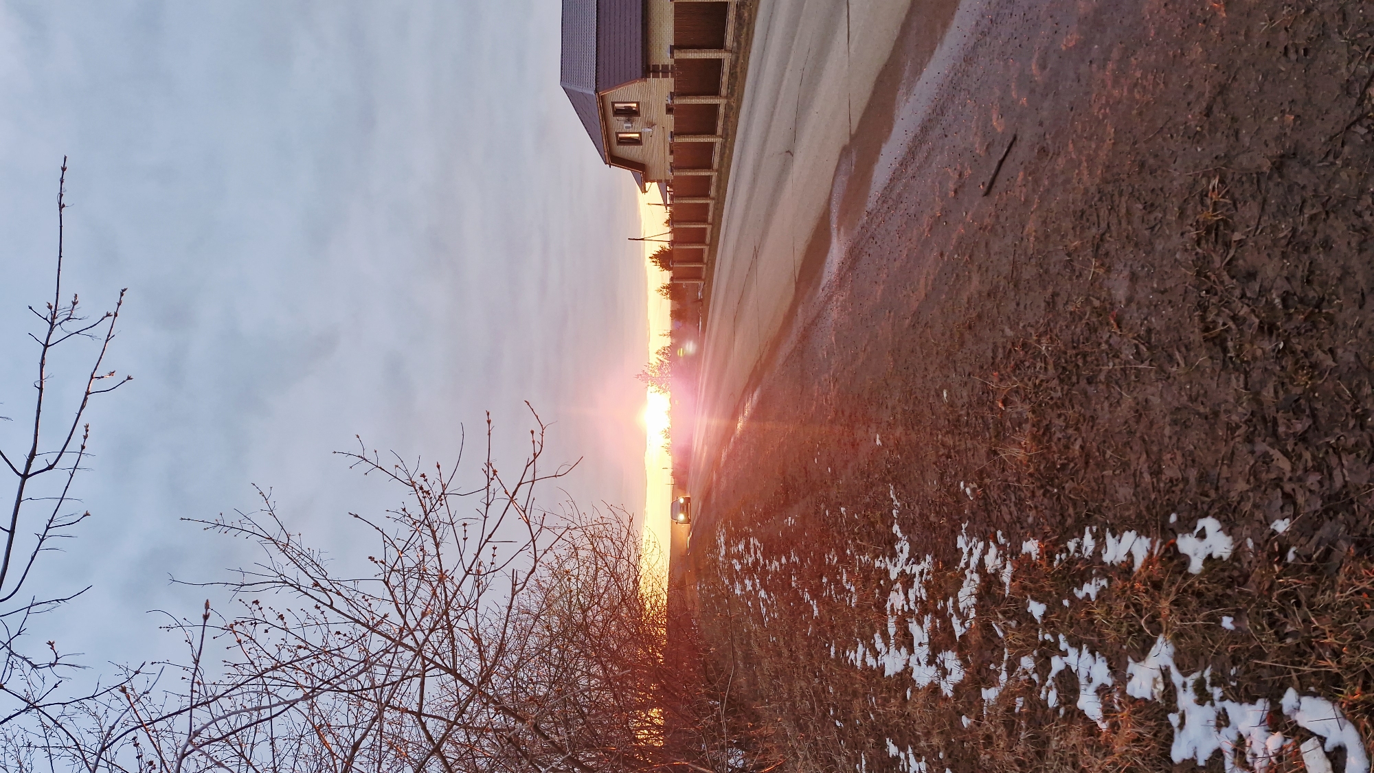 This image, captured in Otchor, Russia, presents a tranquil rural or semi-rural scene during either sunrise or sunset. The sun, a brilliant orange-red orb, is low on the horizon, casting a warm, intense glow across the landscape and creating a distinct lens flare that radiates across the center of the image.
In the foreground on the left, bare tree branches with visible buds indicate a transitional season, likely late winter or early spring. On the right, the ground is a mixture of dark, damp mud, dry leaves, and scattered patches of melting snow.
A paved road stretches into the distance, running parallel to the horizon, with some sections appearing wet. Along the right side of the road, a single-story house with light-colored walls and a dark, possibly tiled, roof stands prominently. It features a series of decorative columns or a fence line along its front. Further down the road, partially obscured by the sun's glare, a vehicle with illuminated headlights is visible.
The sky above is a soft, pale blue-grey, with light clouds, becoming brighter and warmer near the horizon due to the sun's strong light. The overall mood of the scene is calm and serene, underscored by the golden hour lighting and the quiet landscape. There is no visible text in the image.