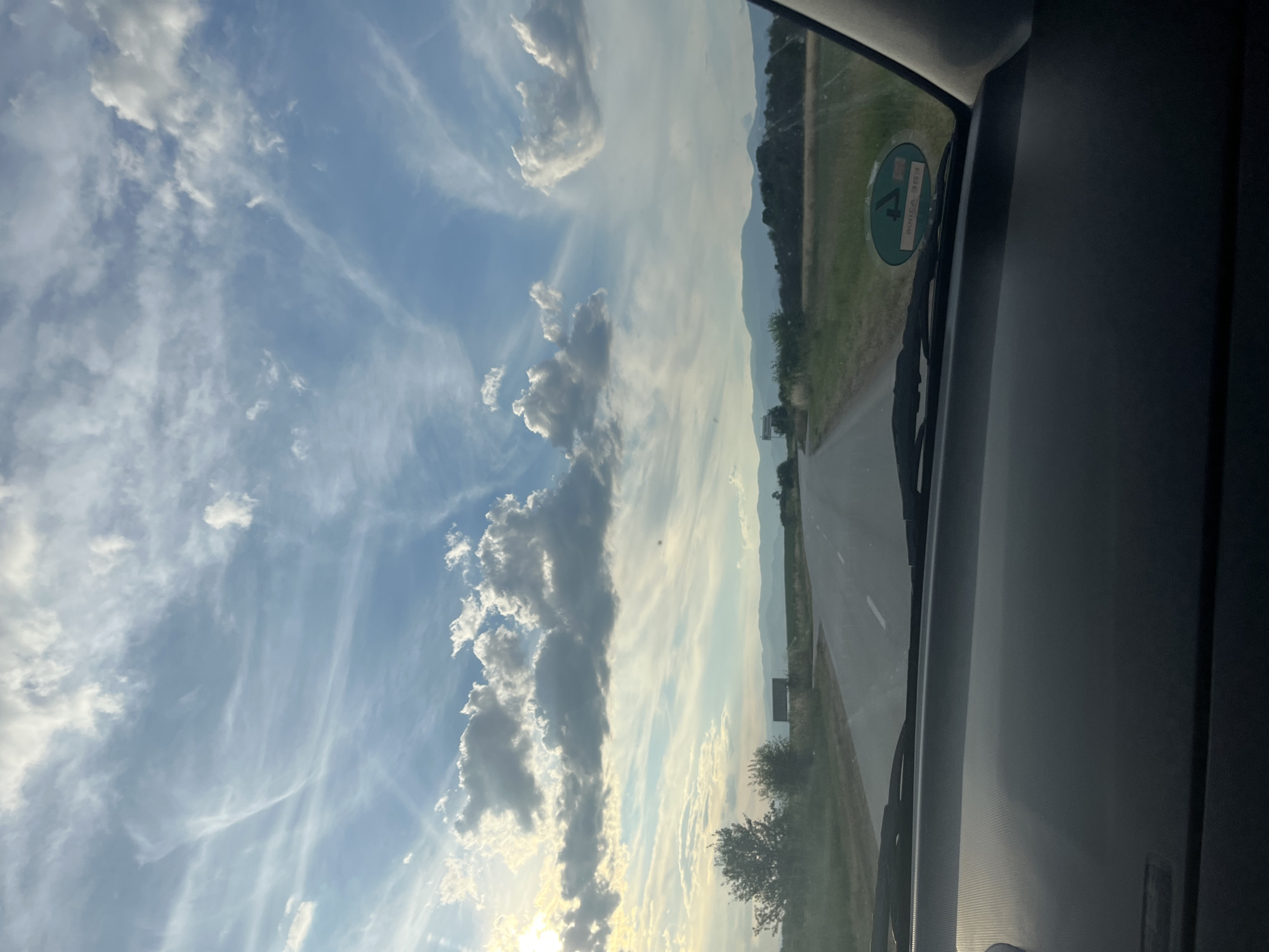 The image captures a view from inside a car, looking out at a rural road in the city of Zhitnitsa, Bulgaria. The sky is a prominent feature, with a mix of blue sky and scattered clouds, some of which are illuminated by the sun. The road ahead is paved and curves slightly to the right, bordered by green fields and trees. In the distance, a range of mountains is visible. The dashboard of the car is visible in the foreground, with a circular green sticker on the windshield on the right side.