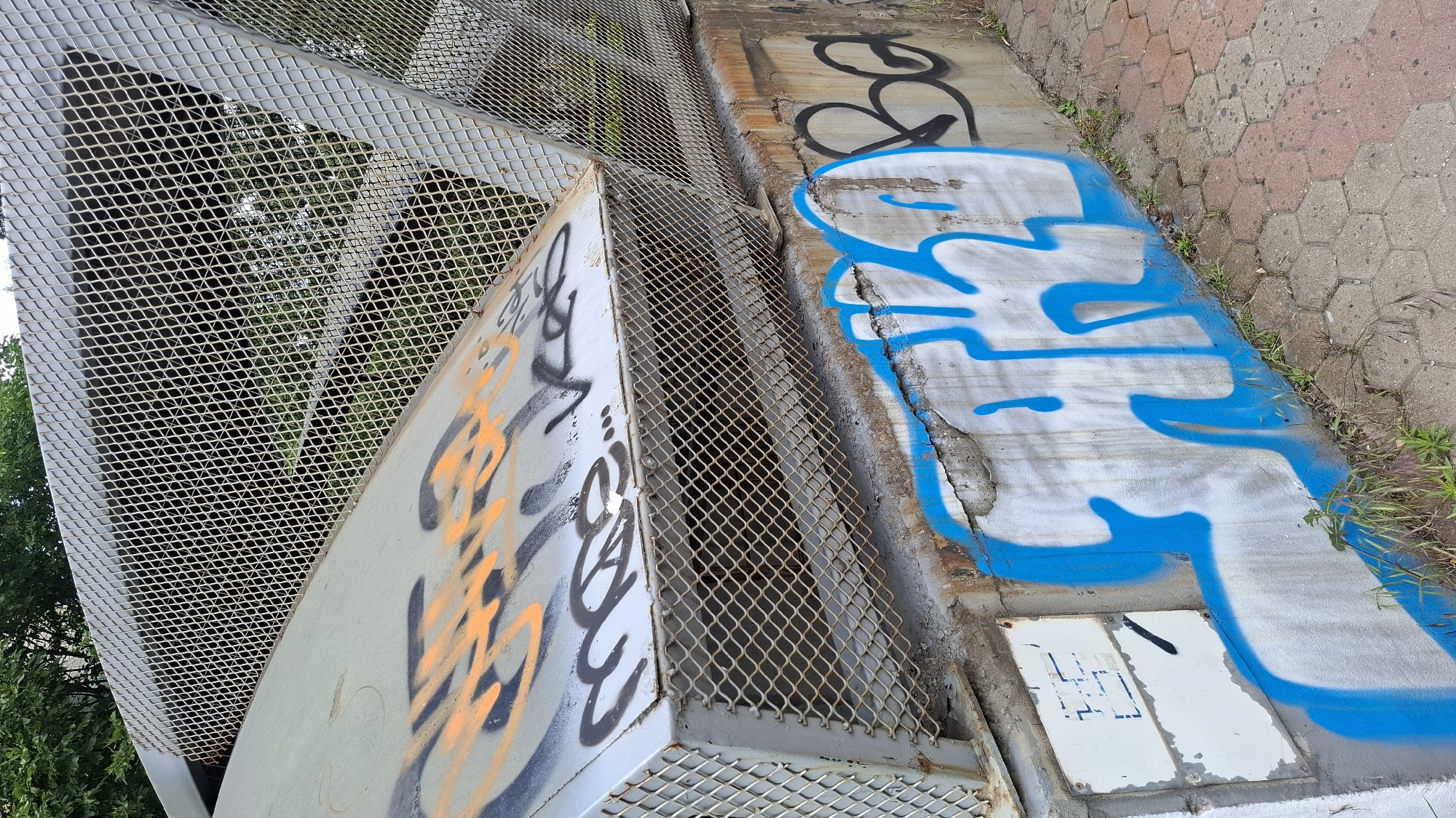 The image shows a section of an outdoor pedestrian overpass or bridge, characterized by a metal grate walkway and a concrete support structure. The concrete surface of the overpass is covered in graffiti, with large blue stylized lettering dominating the right side of the frame and orange and black spray paint visible on a slanted metal panel on the left. The metal panel appears to be part of the overpass's structure, possibly a staircase or ramp.
The scene is outdoors, with greenery and trees visible through the metal grate in the background, suggesting it's a daytime setting. The weather appears to be fair. The ground next to the overpass is paved with hexagonal concrete tiles, some of which are reddish-pink and others gray, indicating an urban or park environment. Small tufts of grass and weeds are growing in the cracks between the pavement stones and along the base of the concrete structure, suggesting a somewhat unkempt or naturalized area.
There is no discernible activity or event taking place; the image focuses on the static elements of the overpass and its graffiti. No people are visible. The graffiti is the most prominent detail, with the blue lettering on the concrete being a large, bold design. The metal grate is a diamond pattern, which is a common industrial design. A small white electrical or utility box is visible on the concrete near the bottom right, with some faint dark markings on its surface.
The text visible is solely graffiti. The large blue spray paint appears to be stylized lettering, but it is not legible as a specific word or phrase. There is also some black spray paint on the concrete to the left of the blue lettering, which is also illegible. On the orange spray-painted section of the metal panel, there are also stylized black and orange markings that do not form legible words. The small white box has some faint blue markings within a square grid pattern, but these are too indistinct to read.
