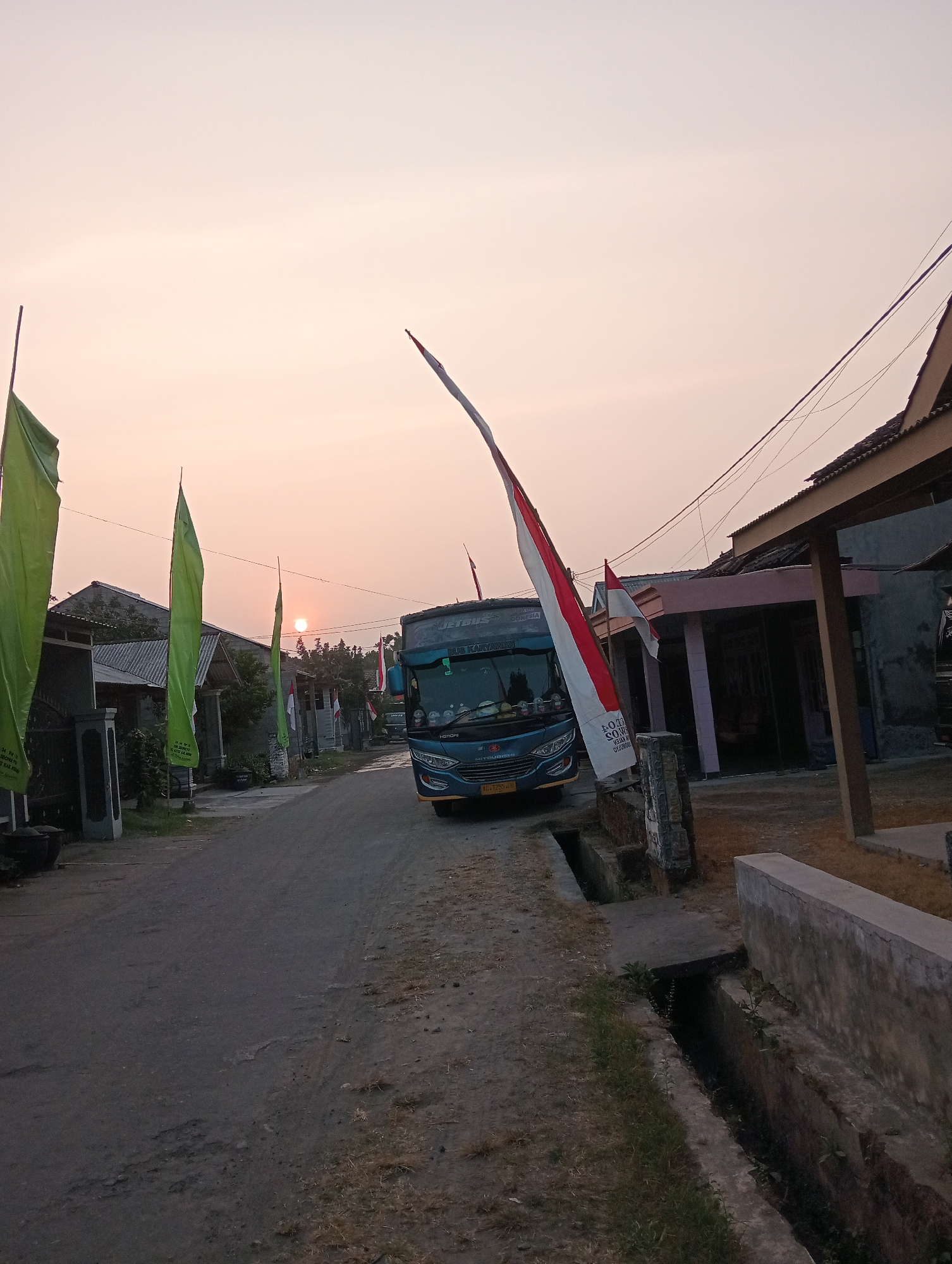 The image depicts a residential street in Wates, Indonesia, during what appears to be sunset. A dark blue bus, identified as a "JETBUS," is parked on the right side of the asphalt road, facing away from the viewer and towards the distance. The bus has yellow license plates. On the left side of the road, several tall, thin flags are visible. Three of these flags are a bright green color, and one is a red and white Indonesian flag, attached to a pole. The Indonesian flag is partially obscuring the front of the bus.
The environment consists of a narrow road with a dirt and grassy shoulder. On either side of the road are houses with tiled roofs and some vegetation. A concrete ditch runs along the right side of the road, adjacent to a low concrete wall. Power lines are strung overhead. The sky is a pale orange and pink hue, indicating the setting sun, which is visible as a bright orb slightly above the horizon, positioned in the distance between the houses. The overall scene suggests a quiet evening in a village or suburban area. There are no people clearly visible in the image.