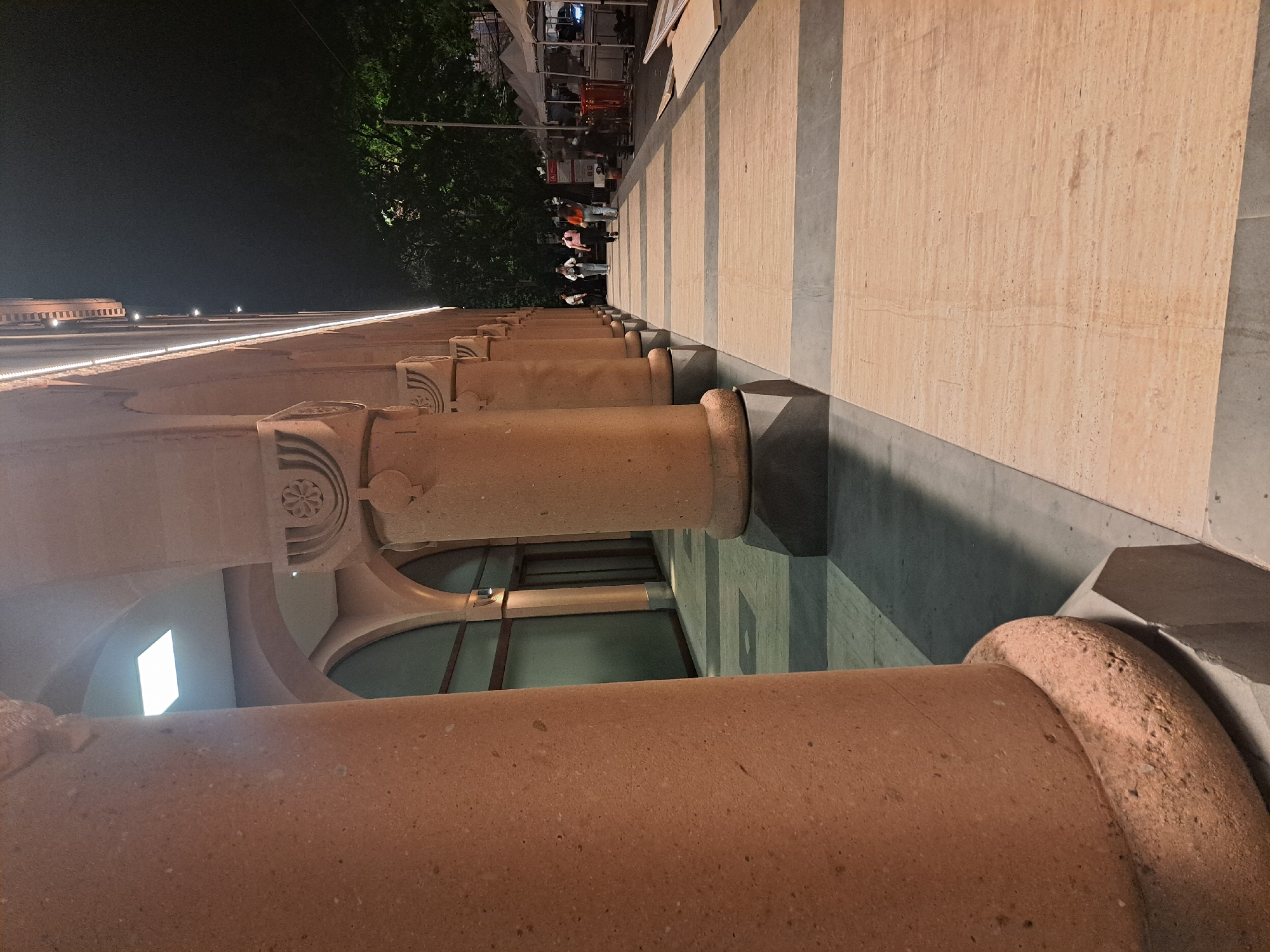This night-time image captures a detailed view of a pedestrian walkway lined by a stone colonnade in Yerevan, Armenia. On the left, a series of large, reddish-brown cylindrical columns with decorative capitals extends into the distance, forming an arcade. The building above the columns features recessed strip lighting along its facade. The paved walkway consists of alternating horizontal sections of light-colored and dark-gray stone. In the mid-ground, approximately five to seven individuals are observed walking away from the camera. To the right of the walkway, dark trees are visible, along with several temporary white-canopied stalls, suggesting a market or event. Construction materials, including wooden planks, are present on the far right. The scene is illuminated by artificial lights, casting shadows on the ground and within the arcade.