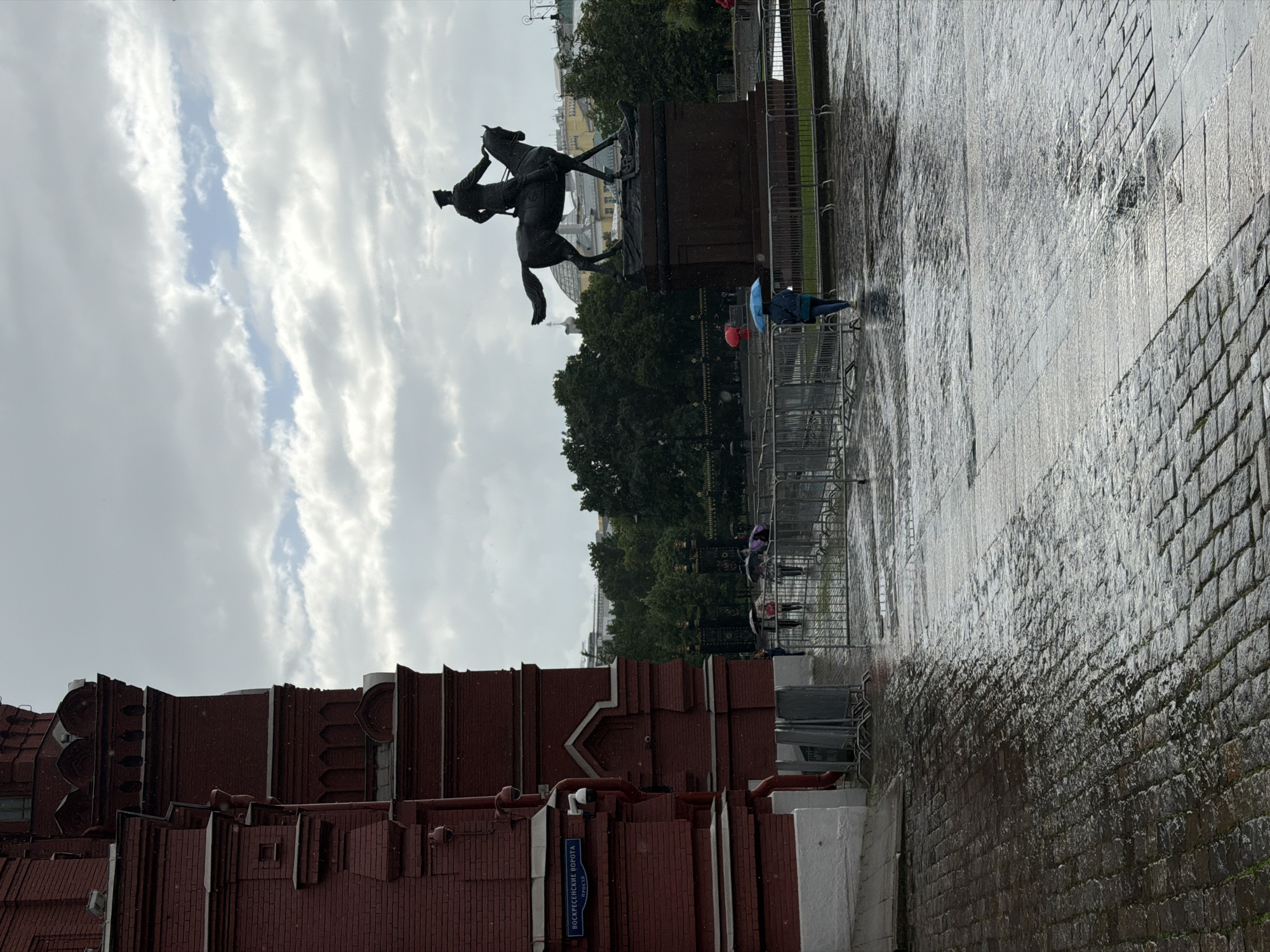 The image depicts a rainy day on the cobblestone pavement of Red Square in Moscow, Russia. In the foreground, the wet cobblestones reflect the overcast sky. A red brick building, likely part of the State Historical Museum, occupies the left side of the frame, with a blue street sign labeled "ВОСКРЕСЕНСКИЕ ВОРОТА" (Resurrection Gate) visible.
Further into the square, a monument to a man on horseback stands on a pedestal. Several people are visible walking with umbrellas, navigating the wet conditions. One person in the mid-ground is using a blue umbrella, while another further away has a red umbrella. Metal barriers are set up across the paved area, likely for crowd control. The background features lush green trees and architectural elements of Moscow. The sky is cloudy with patches of lighter gray and hints of blue, suggesting a break in the rain.