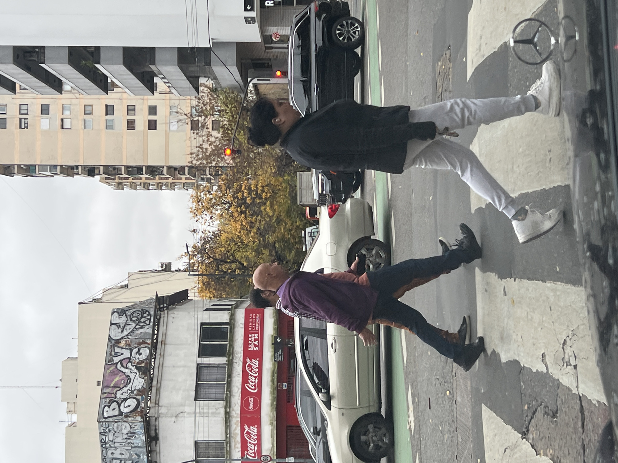 The image captures a street scene in Buenos Aires, Argentina, with two pedestrians crossing a street at a crosswalk. The pedestrians are a bald man wearing a purple jacket and dark pants, and a woman with her hair up, dressed in a black jacket and light-colored pants. They appear to be walking together.
The background reveals elements of the urban environment. A building with graffiti on its side is visible, along with Coca-Cola advertisements on a storefront. Several cars are parked along the street. In the distance, trees with yellowing leaves suggest it may be autumn. Above the pedestrians, a building is seen with stacked balconies and a traffic light. The image seems to have been taken from within a vehicle, as evidenced by a reflection in the window and part of a steering wheel emblem. The image does not contain enough information to pinpoint the exact neighborhood within Buenos Aires.
