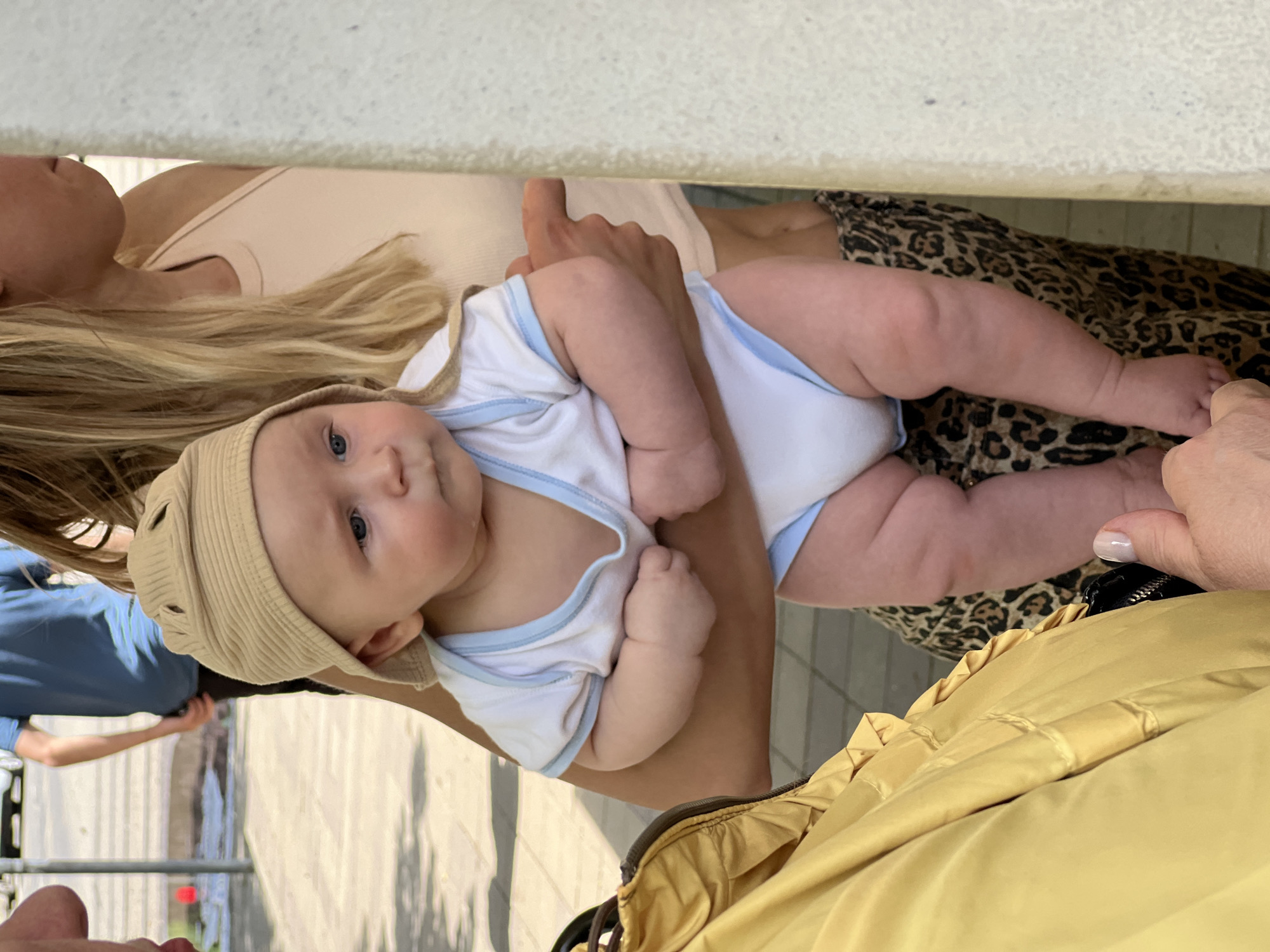 A baby wearing a beige ribbed hat and a white onesie with blue trim is held by a person wearing a tan tank top. The person holding the baby has long blonde hair. The baby is lying on a leopard-print garment. To the right of the leopard print, there is a yellow jacket. Behind the woman is a wrought iron fence, a light-colored walkway, and a blue shirt can be seen in the background. The location may be in the city of Krasnodar, Russia.