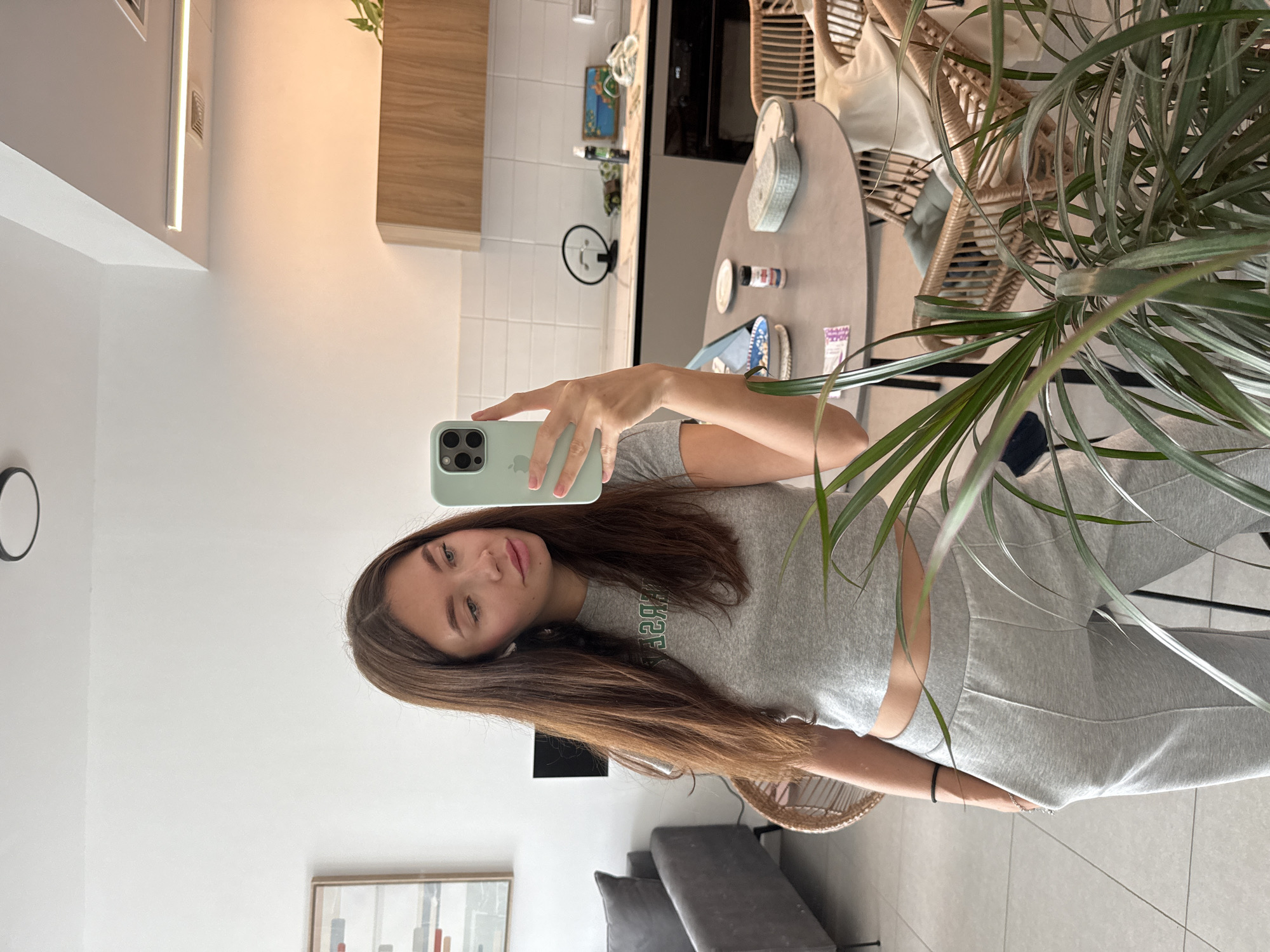 A young woman is taking a selfie in what appears to be an apartment. She is holding a light green phone with three camera lenses. She is wearing a gray t-shirt and sweatpants. Her long brown hair is partially covering the phone. In the background, there is a kitchen area with white tiled walls and a light wood cabinet. A round table with various items is present, along with a wicker chair. A plant is in the foreground. A partial view of a gray couch and an artwork is seen in the distance. There is no definitive evidence to determine that this photo was taken in Ramat Gan, Israel.