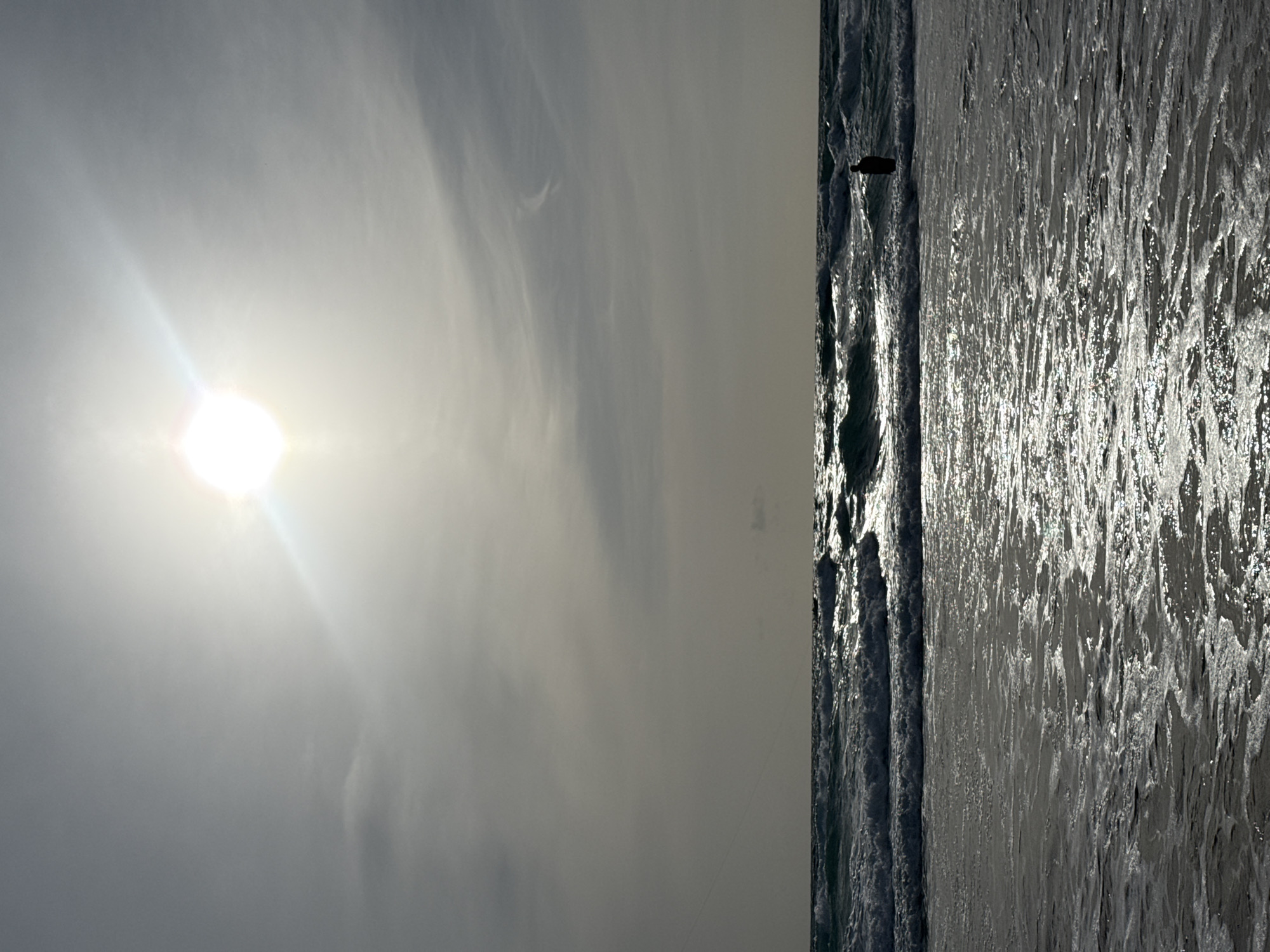 In Karon, Thailand, the sun shines brightly in a hazy sky over the ocean. The sun is high and slightly to the left of center, casting a bright glare across the scene. Waves crash on the shore, creating a frothy, white foam in the foreground. In the distance, a lone figure stands in the water, silhouetted against the bright sea. The scene is serene, with the sound of the waves and the warmth of the sun creating a peaceful atmosphere.