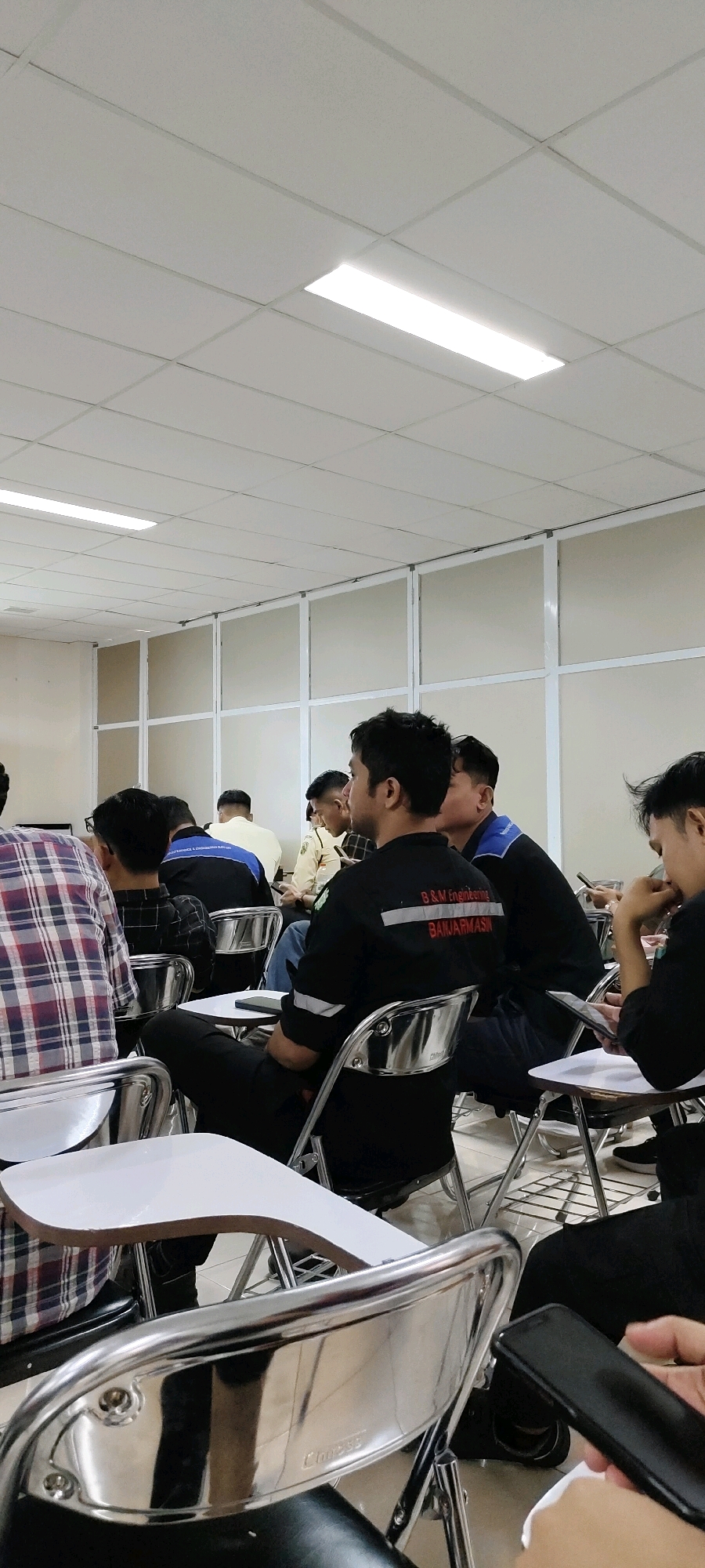 The image depicts an indoor scene, likely a classroom or lecture hall, with a group of young adults seated in chairs, facing away from the camera. The room features a suspended ceiling with fluorescent lighting and partition walls. The individuals are dressed in casual attire, with some wearing shirts that appear to be uniforms or work attire, displaying logos and text such as "B&M Engineering" and "BANJARMASIN". Several individuals are looking forward, suggesting an ongoing presentation or lecture. One person in the foreground is holding a smartphone. The environment is orderly and suggests a formal educational or training setting. The specific location is identified as Nusa Indah, Indonesia.