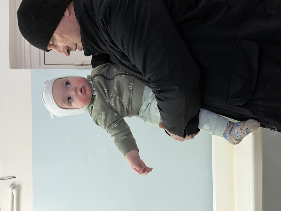 An older man in a black jacket and hat holds a baby. The baby wears a light green jacket with small white figures, light-colored pants with a bear pattern, a matching hat, and patterned socks. The background shows a light blue wall and a white door frame. The man appears to be looking at the baby. The image is rotated slightly, giving the impression that they are tilted.