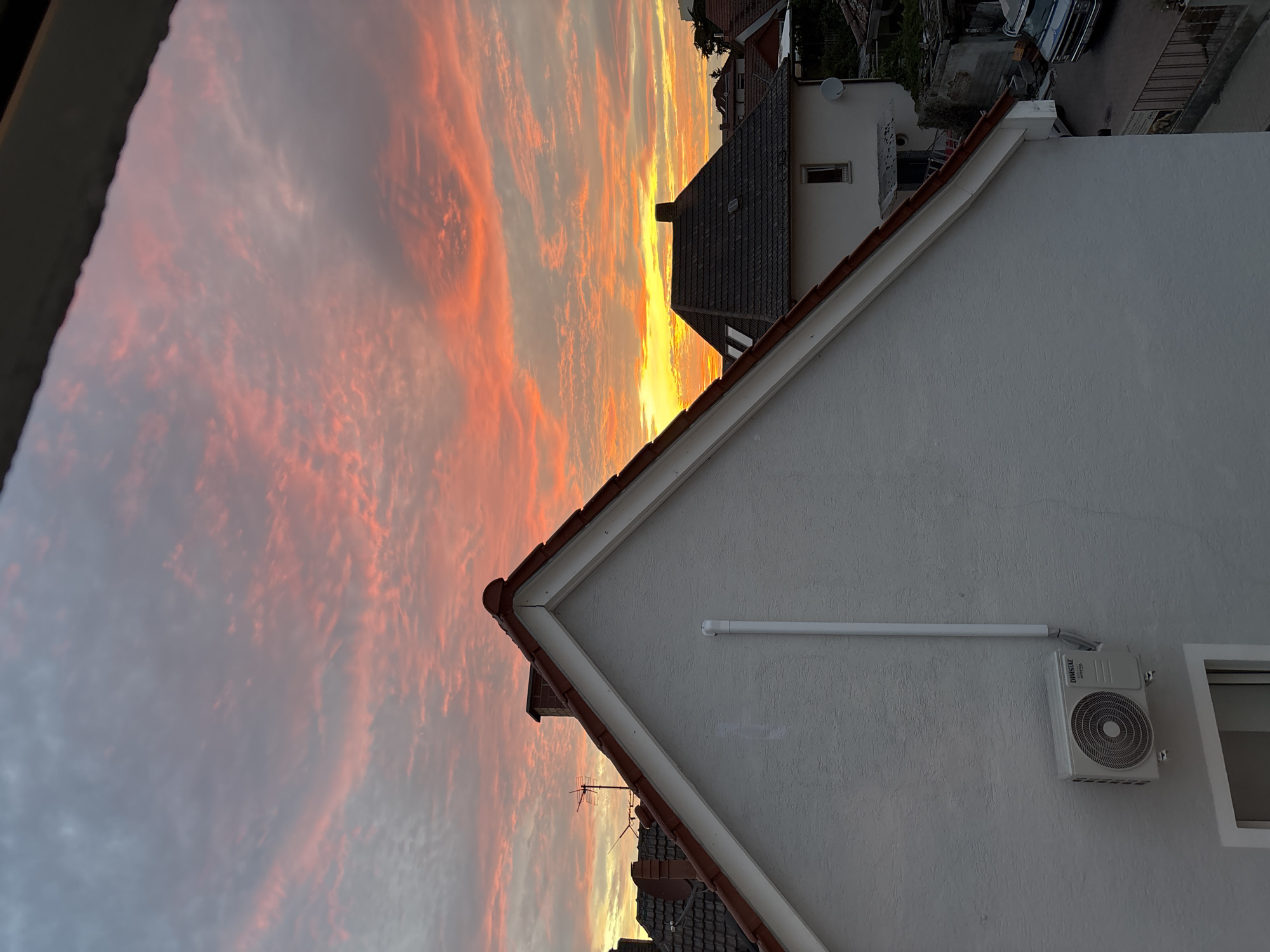 The image depicts a residential area in Viernheim, Germany, under a vibrant sunset sky. In the foreground, a white-painted house with a red-tiled roof dominates the left side of the frame. An outdoor air conditioning unit is mounted on its exterior wall, along with a white pipe. Behind this house, other residential buildings with red and dark grey tiled roofs are visible. A chimney with a red brick top is also present. The sky is filled with dramatic clouds painted in shades of orange, pink, and yellow, with the bright glow of the setting sun visible in the distance. In the background, a street can be seen with a blue truck parked on it and a sidewalk with a metal railing. The overall scene is a tranquil evening view of a suburban neighborhood during a colorful sunset.