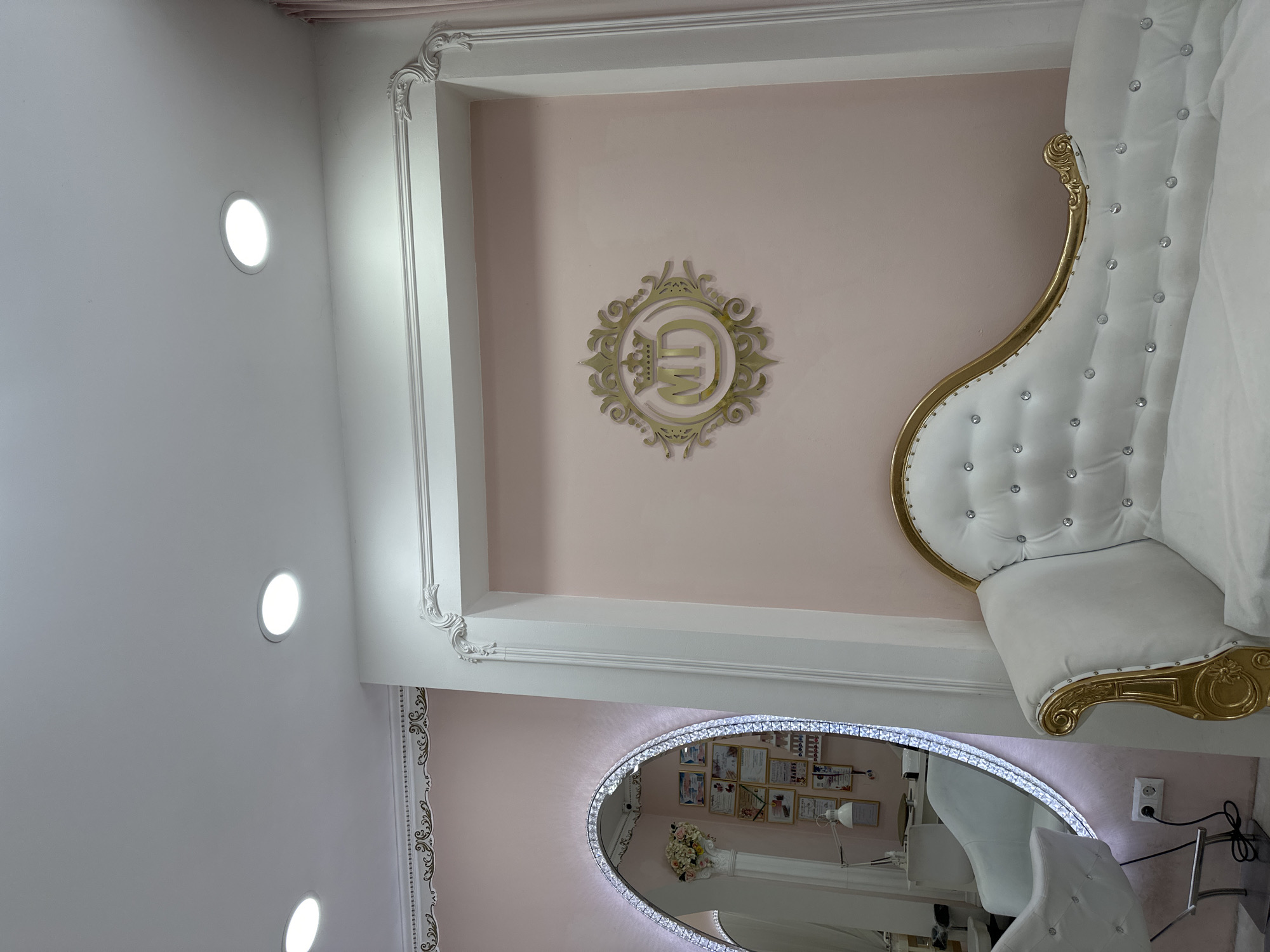 The image shows an interior with a mix of ornate and modern design elements. A white tufted sofa with gold trim is partially visible in the upper right. The walls are light pink with decorative white molding. A round mirror with a crystalline frame reflects the interior, including diplomas, a flower arrangement, and white chairs. The ceiling is white with recessed lighting. There is no information indicating the location is Sofia, Bulgaria. No people or actions are visible in the image.