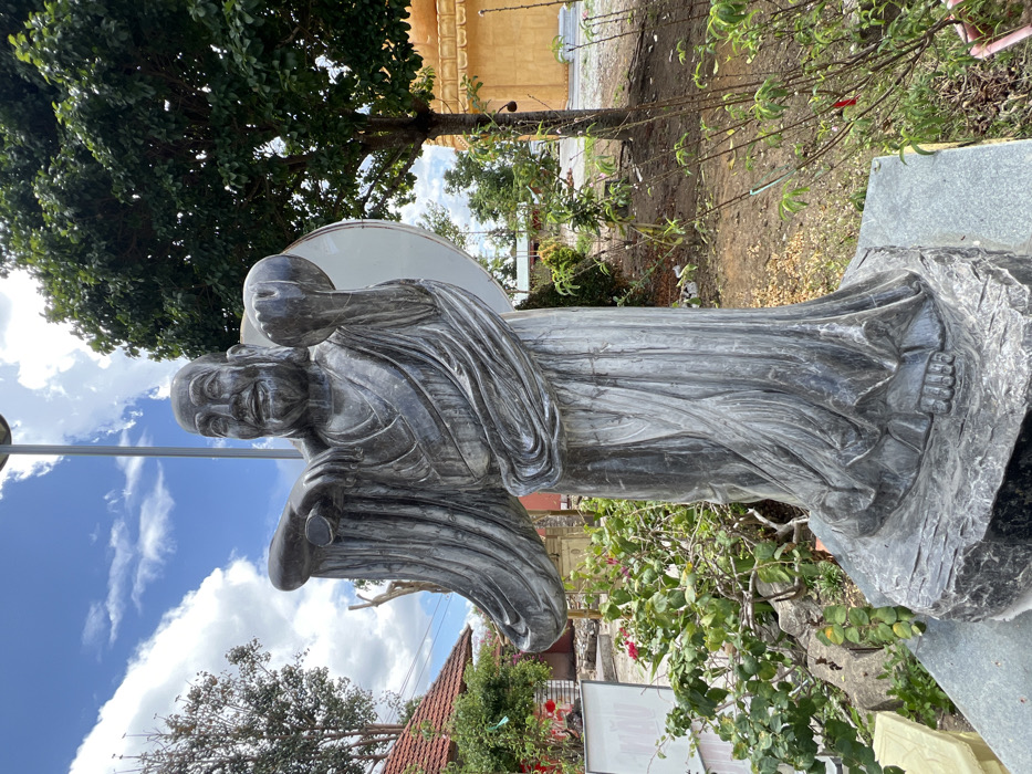 A dark grey stone statue of a smiling, bald, robed figure stands outdoors on a multi-tiered stone base in Đăk Mar, Vietnam. The statue's right arm extends forward, holding a horizontal scroll-like object. Its left arm is bent, holding a large, light-colored disc or circular object behind its back. Green bushes with red flowers are situated to the left of the base, while young plants with green leaves are to the right. In the background, a building with a red tiled roof is partially visible to the upper left. To the right, a large tree with dark green foliage stands, and a golden, multi-tiered religious structure is partially visible behind it. A utility pole extends vertically behind the statue. The sky above is blue with scattered white clouds. The ground is composed of soil and sparse vegetation.