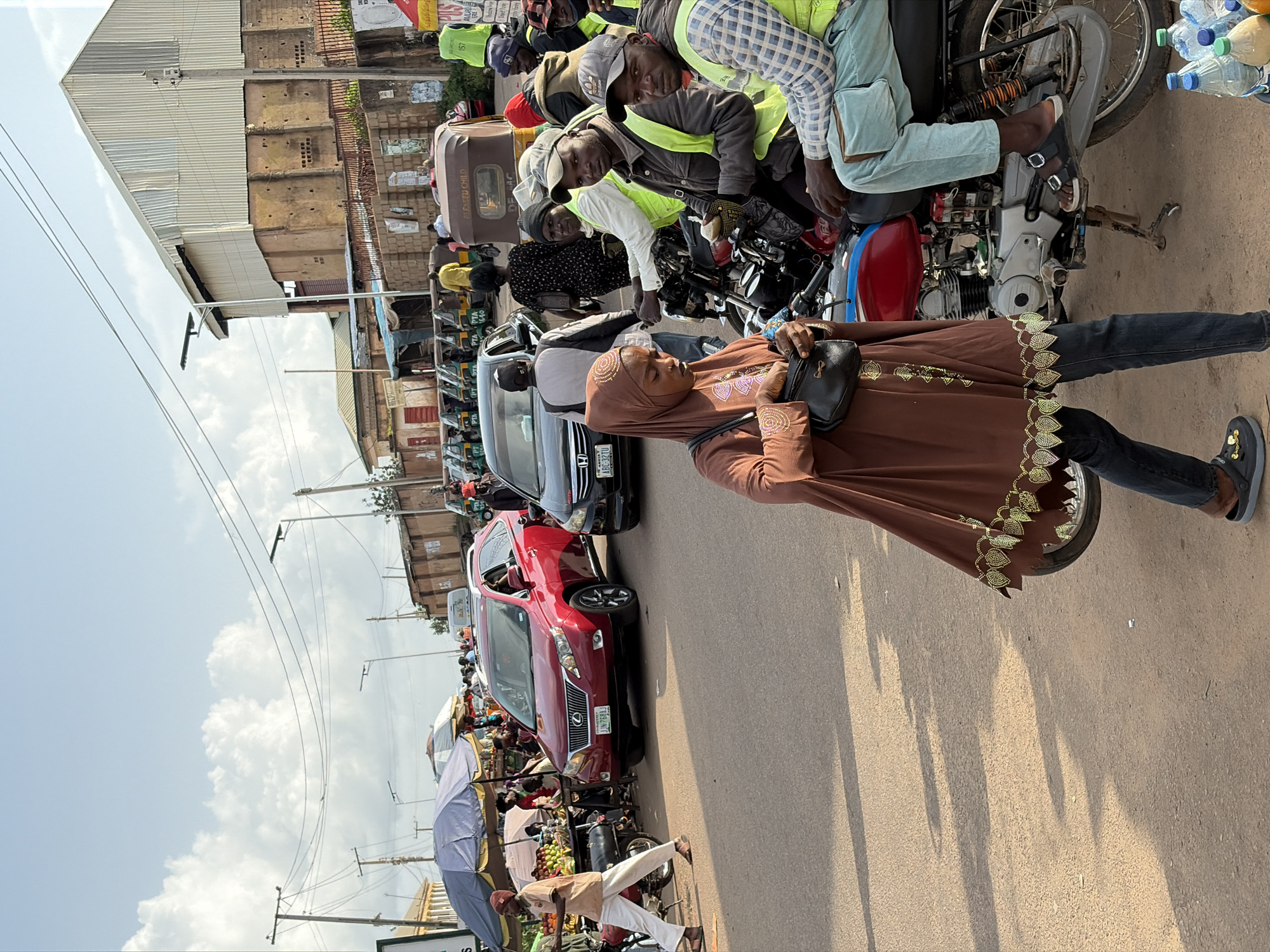In Gura Top, Nigeria, a bustling street scene unfolds under a bright, cloudy sky. A red Lexus SUV is parked on the right side of the road, its passenger-side door open. To its left, a woman in a brown abaya walks away from the camera, holding a black purse. Several men, some wearing bright yellow vests, are gathered on motorcycles. In the background, a line of auto-rickshaws, known as 'Keke Marwa' or 'Keke NAPEP', are parked. Buildings with corrugated metal roofs and brick facades line the street, and overhead, power lines and streetlights are visible. A market stall with blue and white umbrellas and fruit displays is set up on the left side of the road. The road surface is asphalt, with shadows cast by the sun and objects. Bottles of water and other beverages are stacked on the lower right of the frame.