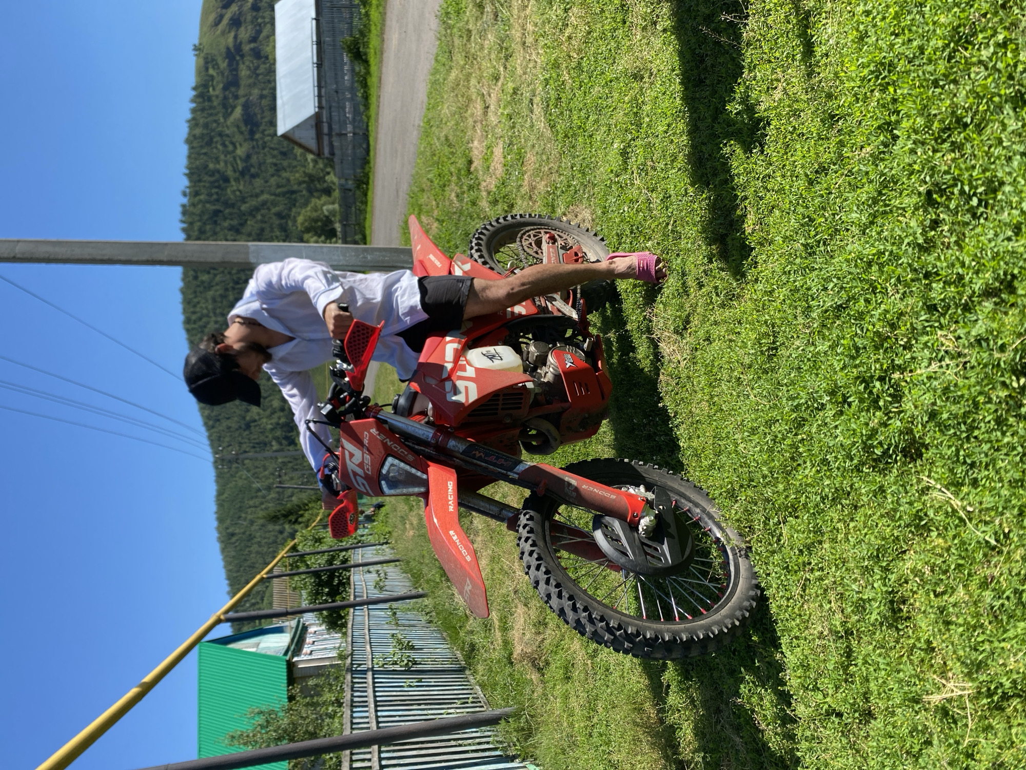 In Maloarslangulovo, Russia, under a clear blue sky on a sunny day, a young man wearing a white long-sleeved shirt, black shorts, a black baseball cap, and pink slip-on shoes is seated on a bright red dirt bike. The motorcycle is parked on a grassy area next to a dirt road. In the background, a wooden fence, a house with a green roof, a power line, and a tree-covered hill are visible. The man is holding the handlebars of the dirt bike, appearing to be about to ride.
