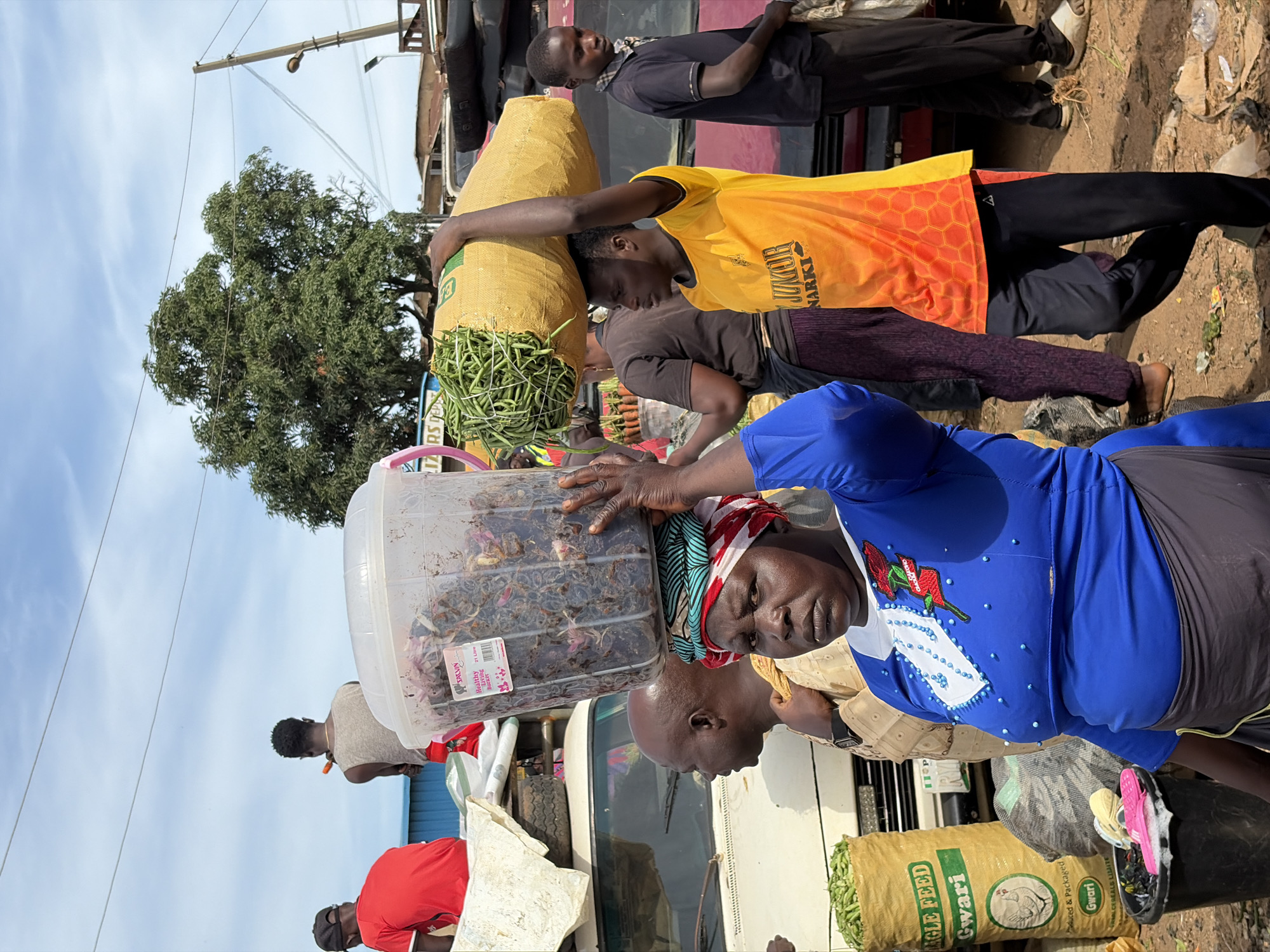 A medium shot captures a busy outdoor marketplace scene in Gura Top, Nigeria, under a bright, cloudy sky. In the foreground, a woman with a headscarf made of red, white, and blue fabric wears a vibrant blue long-sleeved shirt adorned with red floral embroidery and blue beads. She is carrying a large clear plastic container filled with dark, unidentifiable items on her head. To her right, a young man in a bright orange and yellow t-shirt with a hexagonal pattern and black track pants is also carrying a large sack of green beans on his head. Behind them, other individuals are visible, including a man in a light brown garment and another in a black t-shirt and black pants. A yellow sack with the text "SINGLE FEED Gwari" and a chicken illustration is prominent on the left, with a black bucket containing pink flip-flops and white foam nearby. Vehicles, including a white van and a red car, are parked in the background, along with various market stalls and greenery. The ground is dusty, with scattered debris. The overall atmosphere suggests a bustling commerce day.