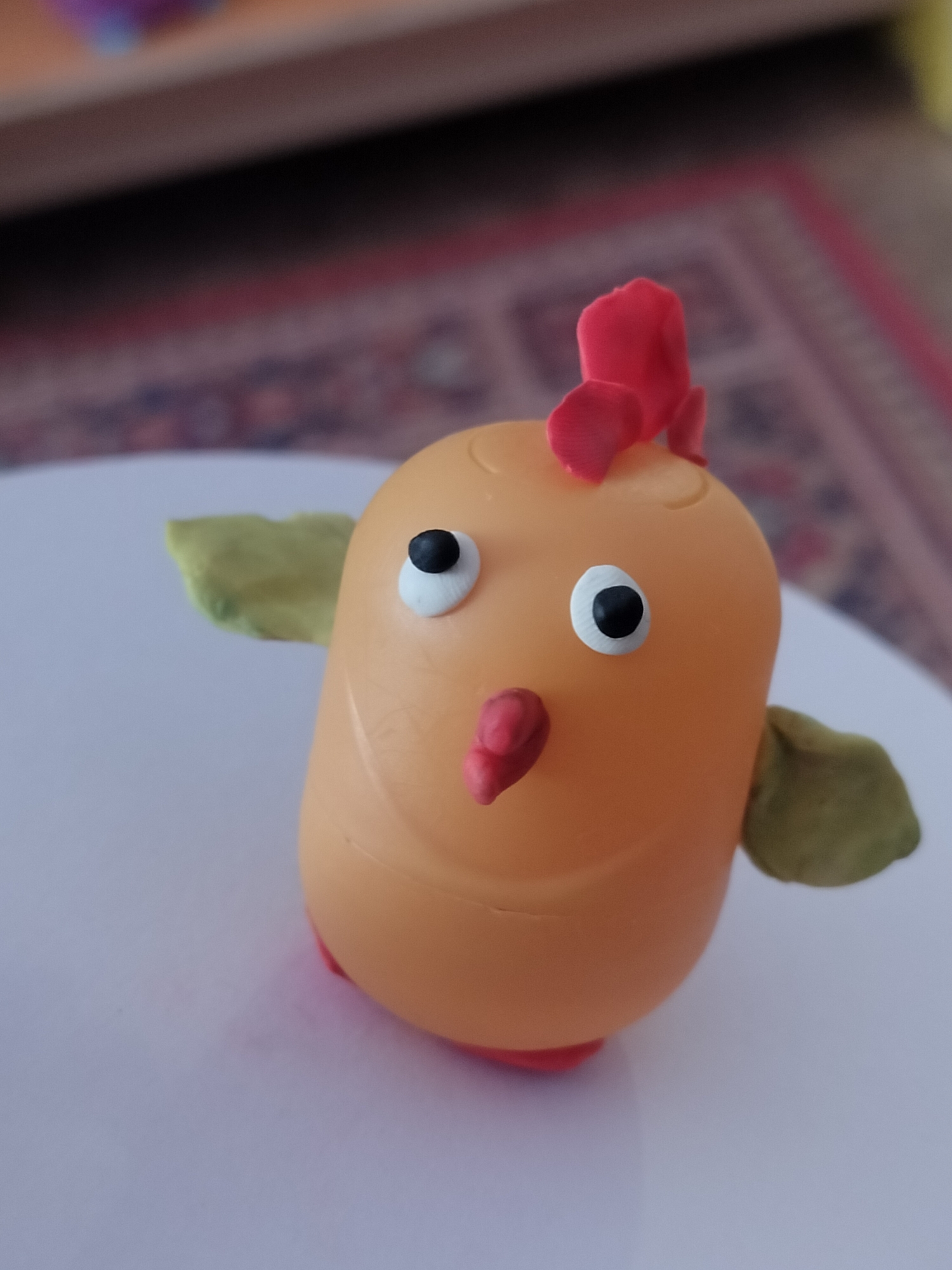A handmade chicken figurine sits on a white surface. The figurine is made from a pale orange, oblong plastic container. It has two googly eyes with black pupils on a white background and a red beak. A red comb is attached to the top of the head. Two green clay-like wings are stuck on either side of the body. The figurine rests on two red clay-like feet. The background is out of focus, but shows a blurred red and brown patterned rug and a dark wooden surface above it. The location and city information given is not possible to determine from this image.