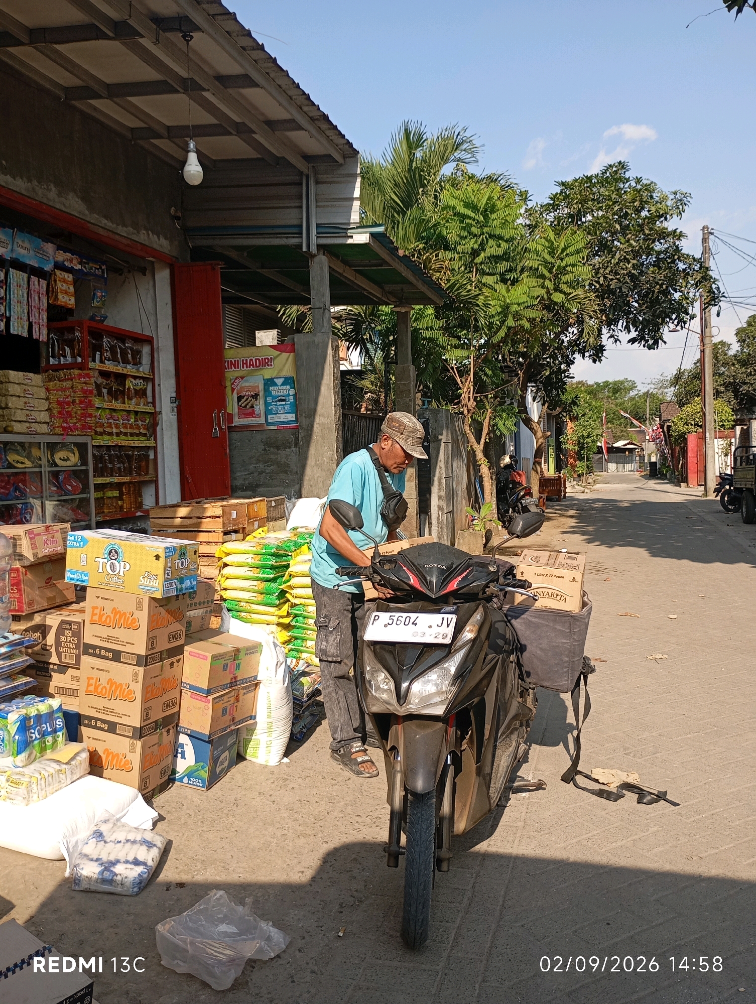 A man wearing a blue t-shirt and a cap is standing next to a black motorcycle. He is reaching towards the motorcycle, which is parked in front of a small store. The store has goods stacked outside, including boxes of food items and bags of rice. The motorcycle has a large storage bag attached to the back. The scene appears to be a street in Rambipuji, Indonesia, with a residential area visible in the background. The day is sunny, and the lighting suggests it is daytime. The man is likely a delivery person or a shop owner interacting with the motorcycle or its contents.