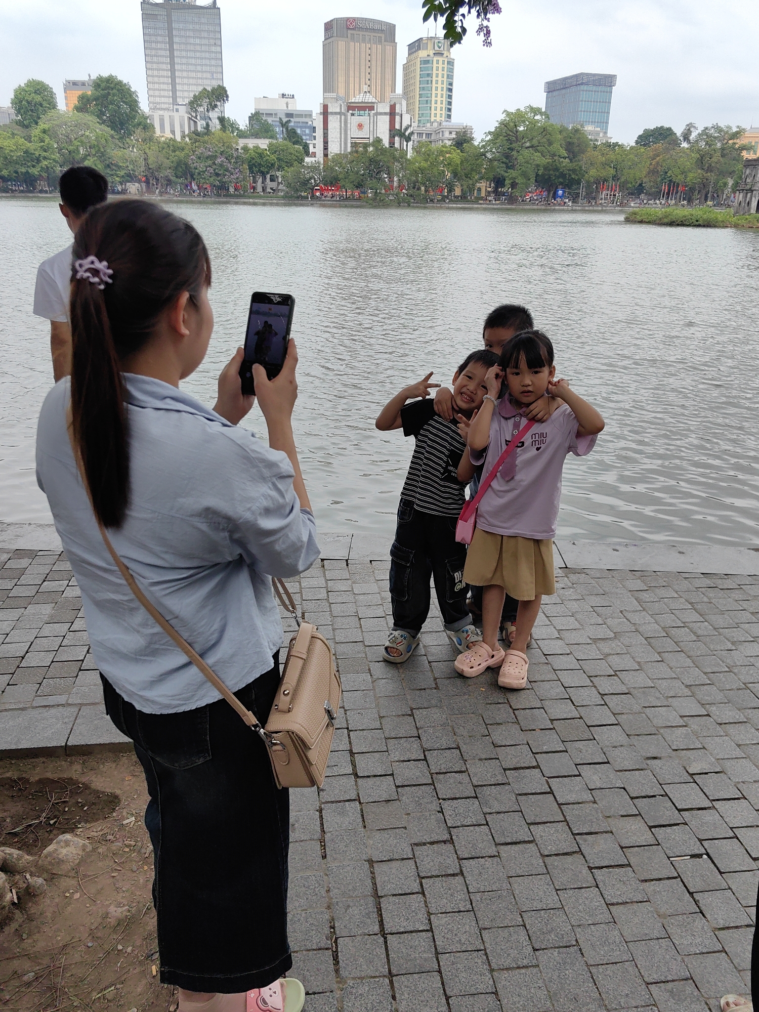 This photograph captures a candid moment by a lake in Hanoi, Vietnam, with a woman taking a picture of three children. In the foreground, viewed from behind, a woman with a ponytail, dressed in a light blue collared shirt and dark skirt, holds a black smartphone to photograph the children. A beige shoulder bag hangs across her. To her left, part of a man's back is visible.
In the mid-ground, three children – two boys and one girl – pose playfully near the water's edge on a grey paved walkway. The boy on the left, in a striped shirt, smiles and gives a peace sign while his arm rests on another boy's shoulder. The girl, wearing a light purple "Miu Miu" polo shirt and a tan skirt with a pink strap, looks at the camera with a neutral expression, hands near her ears. All children wear sandals, including pink Crocs on the girl.
The background features a calm lake, its surface gently rippled under an overcast sky. The far shore is lined with trees and several modern high-rise buildings. The scene suggests a daytime family outing.