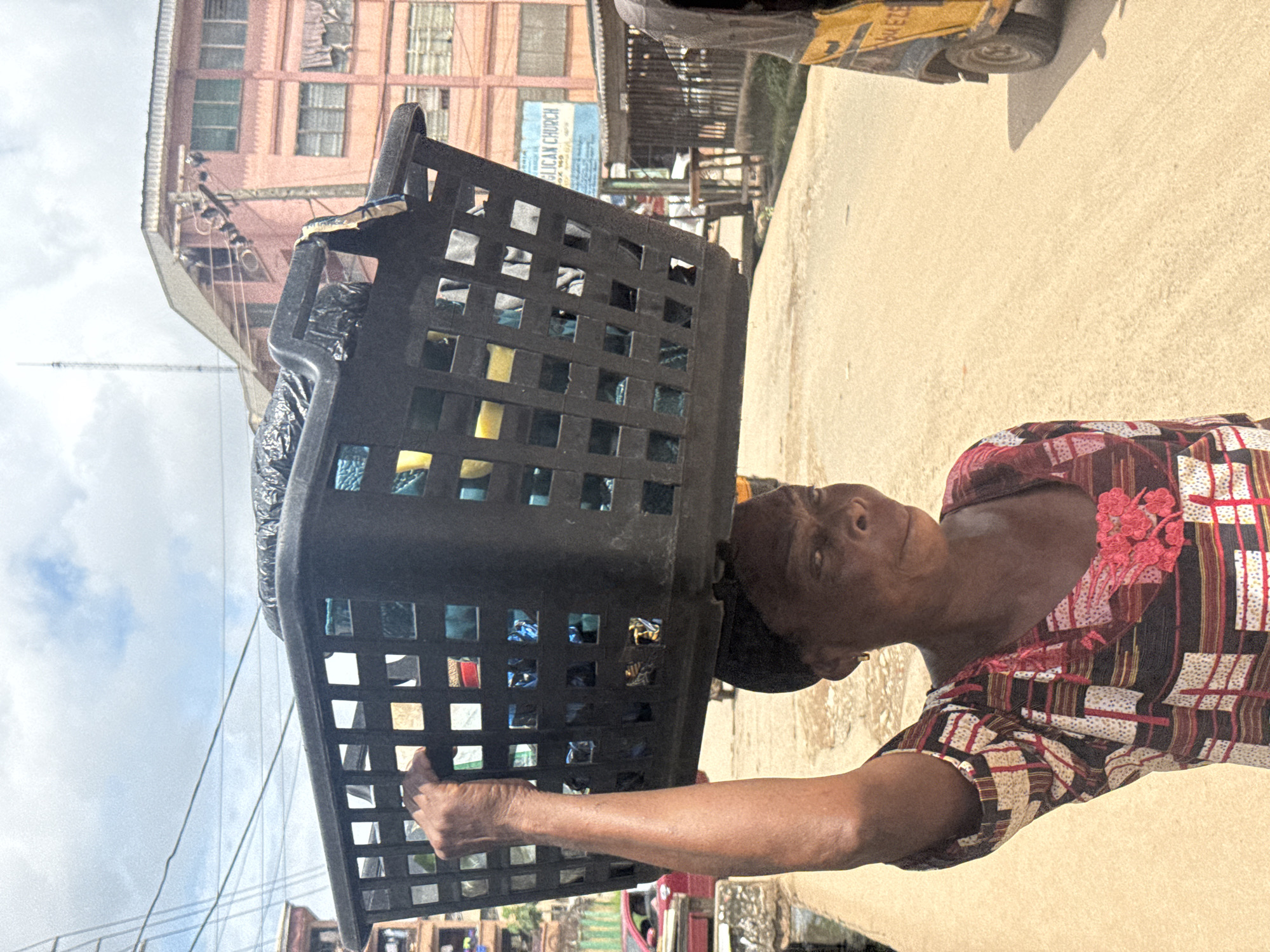 A woman carries a large black plastic basket filled with items on her head. She is walking on a dusty street with buildings lining the background. The woman is wearing a colorful patterned dress. To her right, a yellow motorized rickshaw is partially visible. In the background, a sign for "APOSTOLIC CHURCH" is displayed on one of the buildings. The sky is cloudy.