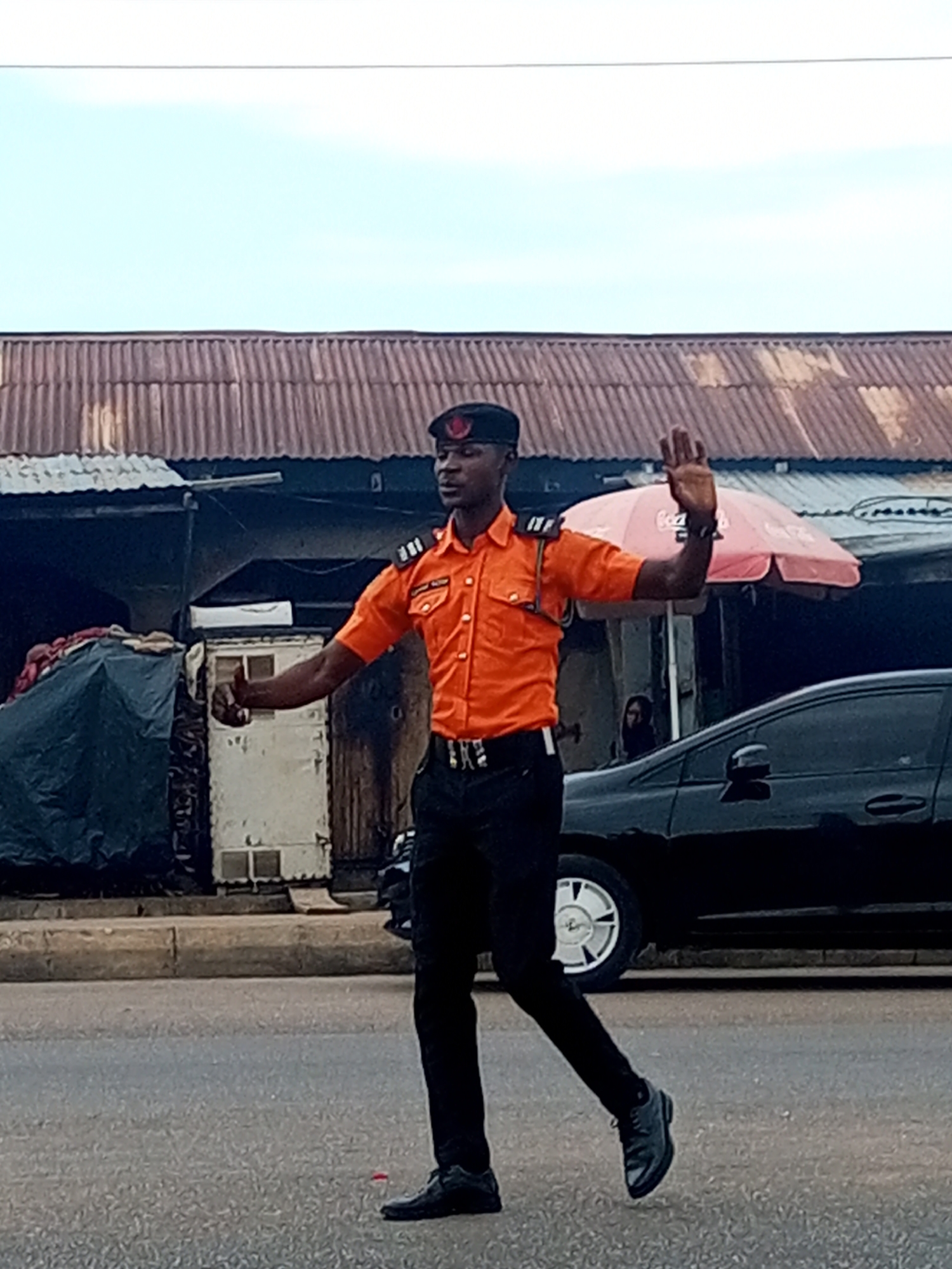 This forensic description details an urban street scene featuring a man in uniform.
**Location:** Unknown urban street in the city of Unknown, Unknown.
**Description:**
The image shows a dark-skinned man, approximately mid-20s to mid-30s, standing on a dry, dark grey asphalt road, actively directing traffic. He wears an orange short-sleeved collared shirt with epaulets displaying three white horizontal bars, black trousers, black lace-up shoes, and a black beret adorned with a red emblem on the front. A nametag on his right chest is partially legible, indicating "COMMUNITY." His right arm is raised and bent at the elbow, with his hand open, palm facing forward and fingers extended, signaling a stop. His left arm is extended horizontally outwards, palm down, as if gesturing for movement or direction.
The background consists of an array of urban structures. Across the mid-ground, a line of buildings is topped with aged, corrugated metal roofs. To the man's left, a dark-colored, possibly black, tarp covers a large object or stall, adjacent to a lighter-colored, possibly white, small building featuring small rectangular windows. To his right, a red and white umbrella, partially branded with "Coca-Cola," suggests a vendor stall, with a dimly visible person standing behind it. A black sedan is visible in the right mid-ground, parked or moving slowly alongside a concrete curb that delineates the road. The sky overhead is a clear, light blue, indicating daylight conditions.