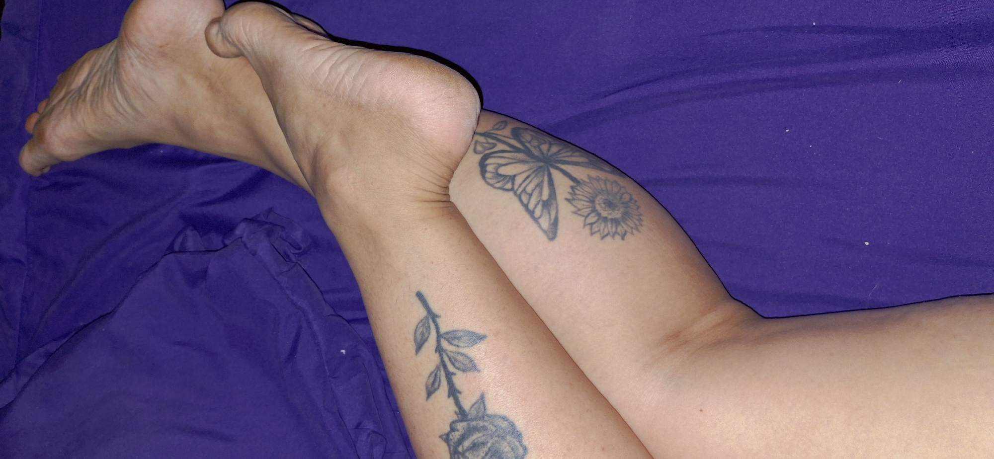 The image shows a close-up of a person's legs and feet resting on a purple fabric. The legs have tattoos, including a rose on the left leg and a butterfly with a sunflower on the right leg. The person's feet are visible, with the soles facing upwards. The fabric appears to be a blanket or sheet, and it is draped loosely, creating folds and shadows. The lighting is soft, and there are some small white specks on the fabric that could be lint or dust. The image is taken from a slightly elevated angle, looking down at the legs and feet. There is no information in the image that places it in Vallecito, Venezuela, or any specific city.