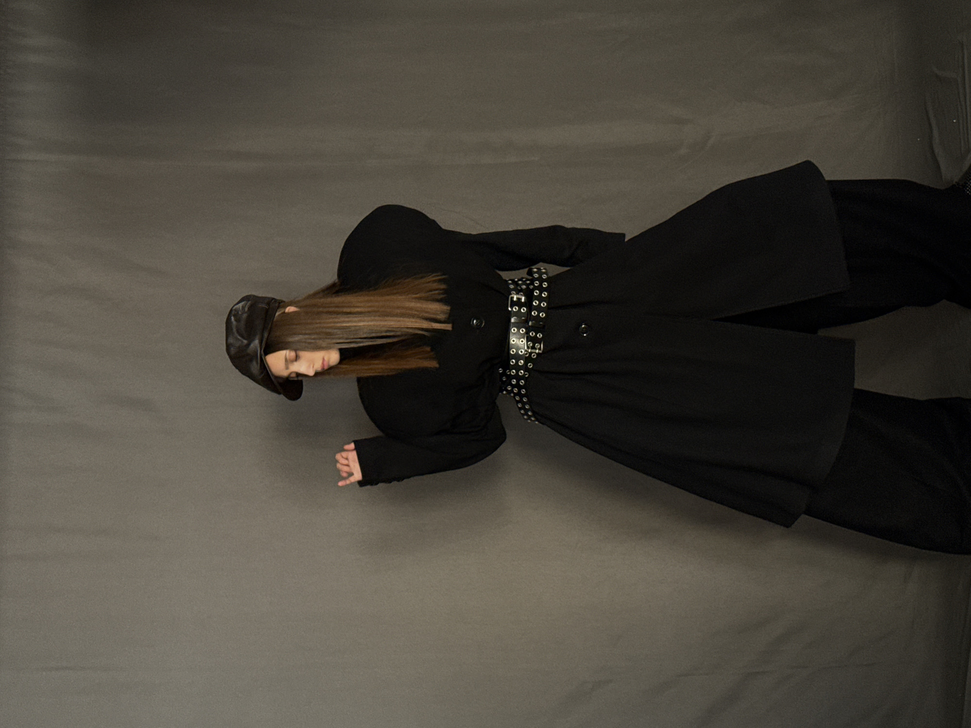 A person is lying on a gray surface, angled toward the upper left corner of the frame. They are wearing a black leather cap, a black coat belted with a silver-studded belt, and black pants. The person's hair is light brown and straight. The background is a uniform gray. No specific features or details suggest the location is in the city of London, United Kingdom.