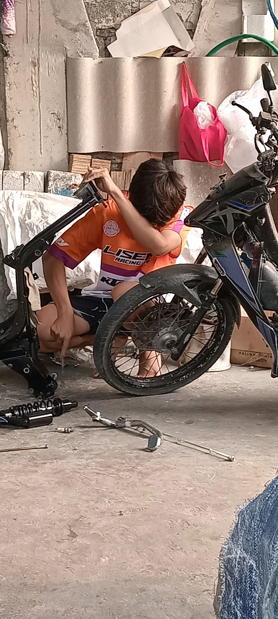 A young male is working on a disassembled motorcycle frame in an outdoor area that appears to be a residential yard or workshop. He is wearing an orange t-shirt with "LISEN RACING" printed on it and dark shorts. The motorcycle frame is black, and some of its components, including a shock absorber and part of the exhaust, are on the concrete ground. The background shows a rough, textured wall, possibly made of concrete blocks, with some discarded materials, including white plastic sheeting and a pink bag. The scene suggests a repair or assembly process taking place in a semi-organized, possibly low-income environment. The overall lighting indicates it is daytime. The location is identified as Babat, Indonesia.