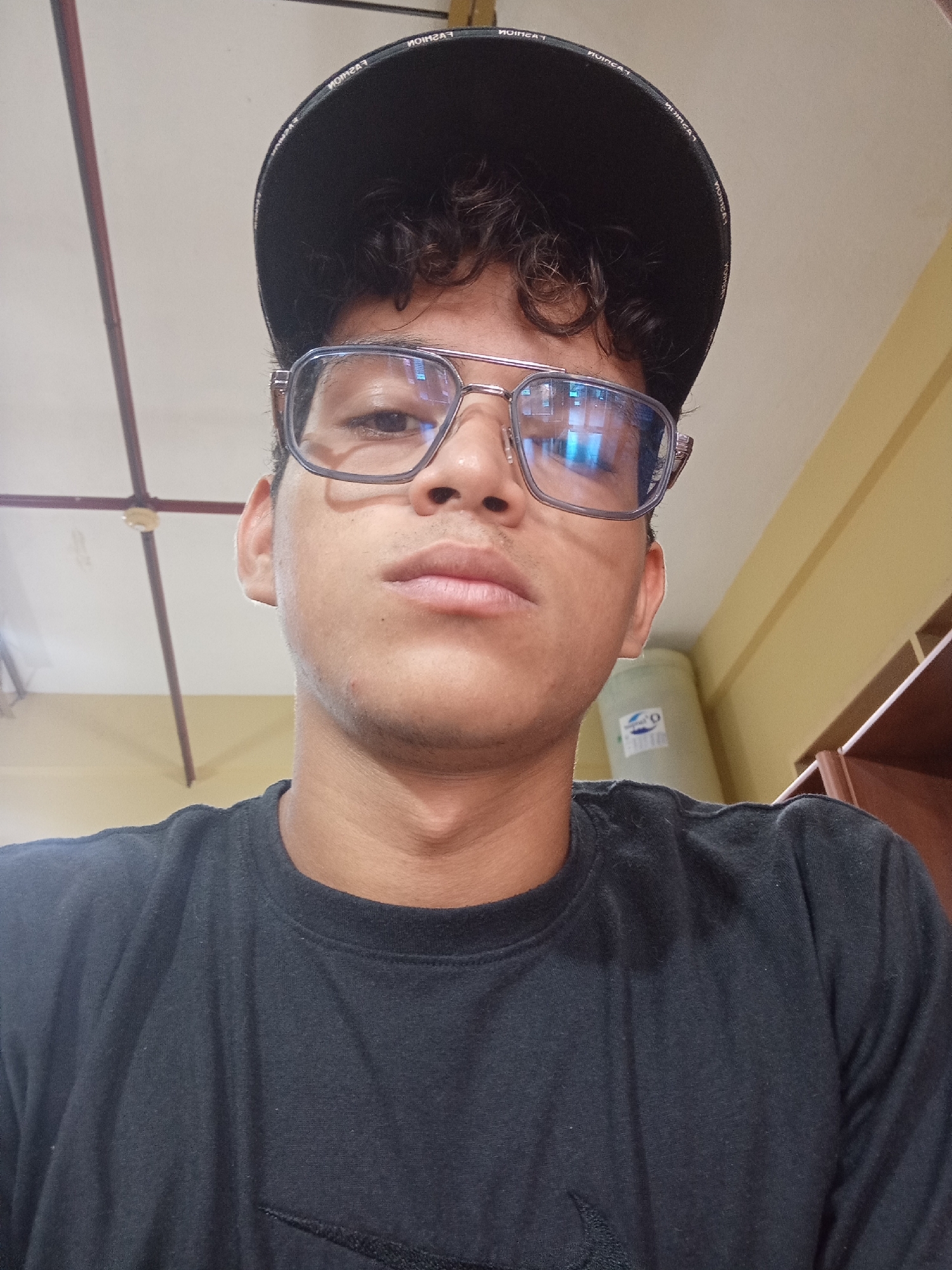 The image is a close-up, waist-up selfie of a young person wearing a black t-shirt and a black baseball cap. The person is also wearing rectangular eyeglasses with a metal frame. The background appears to be an indoor setting with a light-colored ceiling and walls, and a ceiling fan visible. A light-colored cylindrical object, possibly a water tank, is also in the background. The person is looking directly at the camera. The location is identified as Juan Griego, Venezuela.
