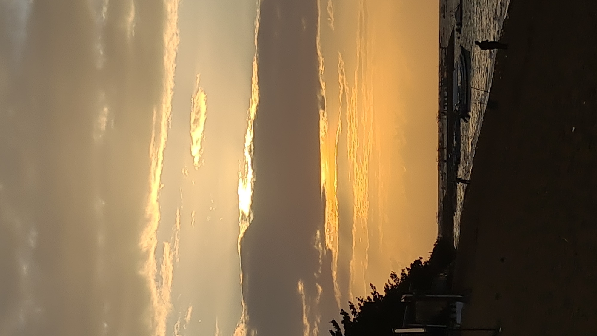 This media file captures a sunset over the ocean, likely in Denpasar, Indonesia, given the provided location context. The sky is dominated by warm, golden hues, with clouds creating dramatic light and shadow patterns. A sliver of sunlight breaks through the clouds, illuminating the scene with a soft, ethereal glow.
The foreground features a beach area, partially obscured by darkness. Several objects are visible on the beach, including what appears to be a traditional fishing boat. A few silhouetted figures can be seen near the boat and along the shoreline, suggesting a tranquil evening atmosphere. The water's surface reflects the warm colors of the sunset, adding to the serene ambiance. The overall mood is peaceful and contemplative, evoking the beauty of a tropical sunset. The time of day is clearly sunset, and the weather appears clear with scattered clouds.