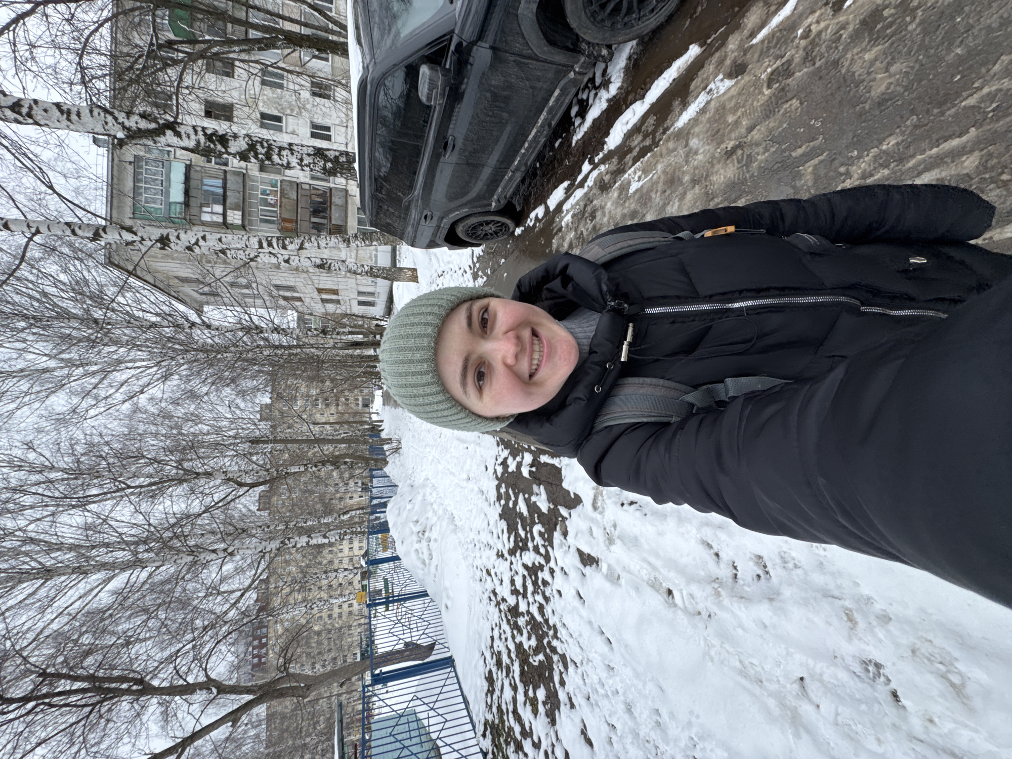 A person is standing on a snowy sidewalk in what appears to be the city of Izhevsk, Russia. The person is wearing a black winter jacket, a light green beanie, and a backpack. There is a black car parked on the side of the street, and a building can be seen in the background. Trees with bare branches are visible along the side of the street, and a metal fence runs alongside the sidewalk. The ground is covered in snow and dirt.