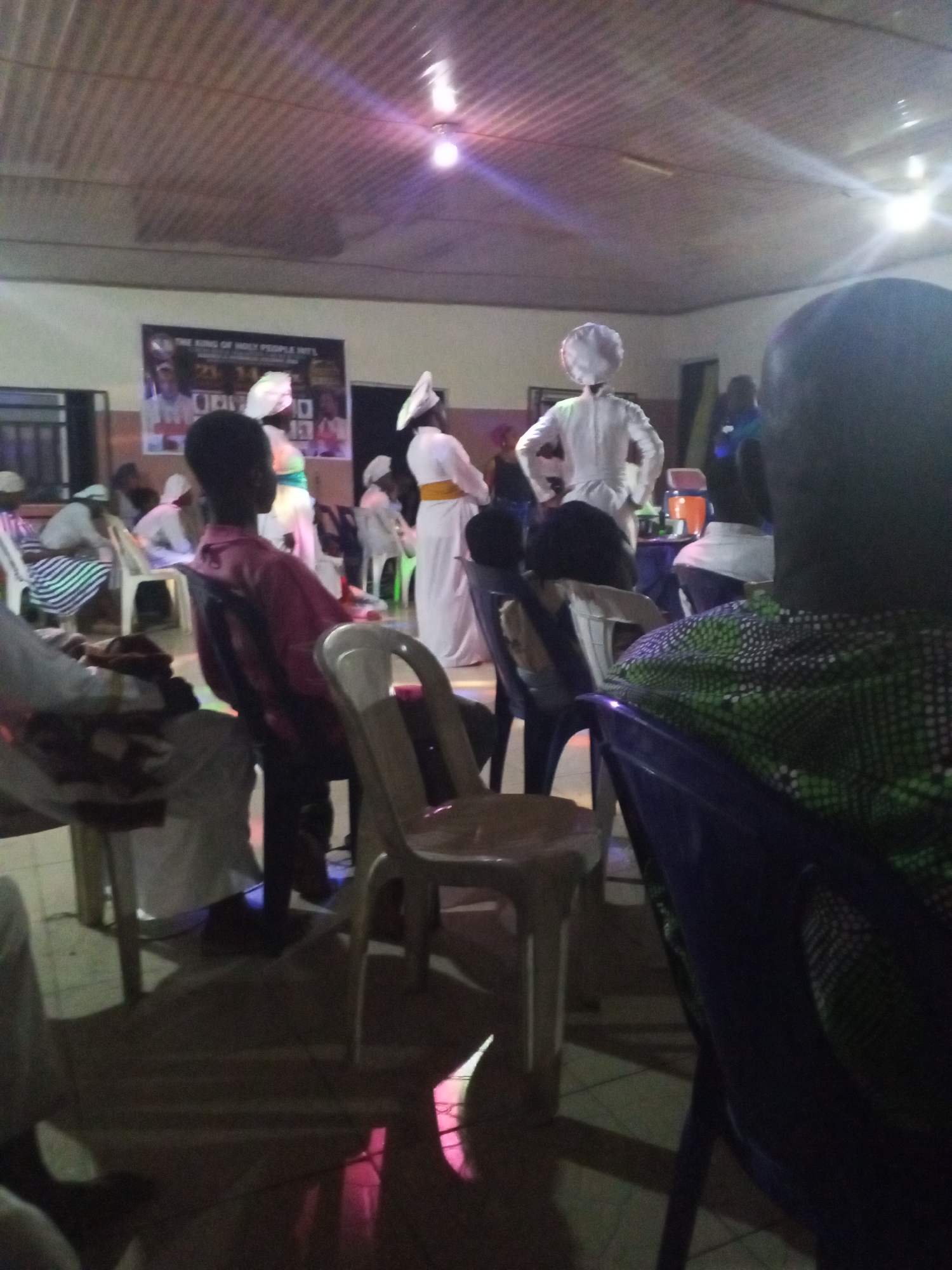 The image captures a scene inside what appears to be a religious gathering in Lokoja, Nigeria. The location is a room with a metal ceiling and fluorescent lighting. There are several people seated in plastic chairs, facing a group of individuals dressed in white robes with head coverings, some with yellow sashes. The people in white robes seem to be performing or presenting something. There is a poster on the wall behind them. A refreshment table with a drink dispenser is visible to the right of the group in white robes. The overall impression is that of a church or religious service.