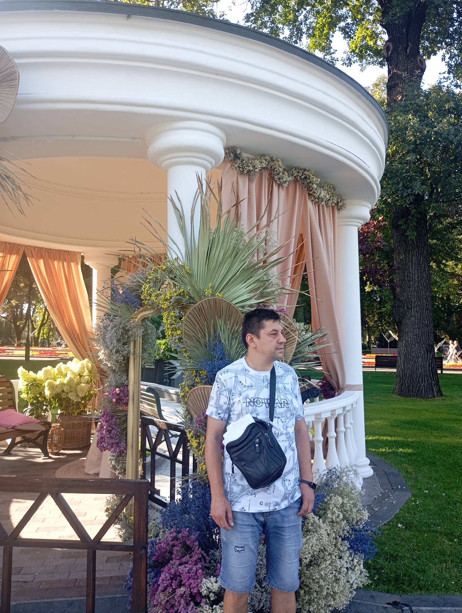 A man stands in front of a white rotunda decorated with dried flowers and palm leaves in a park. The man, wearing a white t-shirt, denim shorts, and a black crossbody bag, looks to his right. The rotunda has a white railing and a peach-colored curtain. The background features a large tree, green grass, and a flower bed. The scene appears to be in Kharkiv, Ukraine.