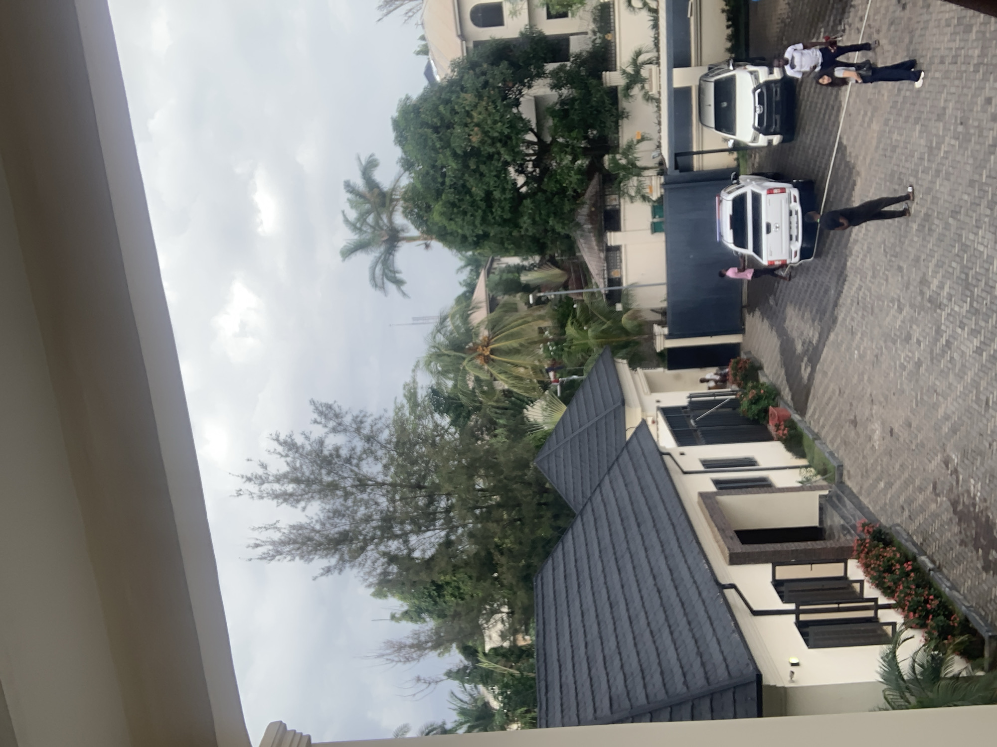 The image depicts an exterior view of a residential property, possibly in Abuja, Nigeria, on an overcast day. In the foreground, a portion of a building's overhang is visible, framing the scene. The property features two modern-style houses with dark-tiled roofs and light-colored walls. Flowerbeds with pink and red flowers line the walkways and the front of the houses.
A paved driveway and courtyard are visible, with two white vehicles parked: a pickup truck with emergency lights on top and a larger SUV. Several individuals are present in the courtyard. One person in a pink shirt is near the pickup truck, while another in a black t-shirt and white shorts is walking towards the pickup truck. Two other individuals, a woman in a grey top and dark pants, and a man in a black and white striped shirt, are standing near the SUV. A person in a black t-shirt and dark pants is also standing near the pickup truck, facing away from the camera.
Behind the houses, lush green trees and palm trees are abundant, suggesting a tropical or sub-tropical climate. In the background, larger multi-story buildings are visible, hinting at a suburban or urban residential area. The sky is cloudy, casting a soft, diffused light over the scene. The overall impression is of a well-maintained, affluent residential setting.
