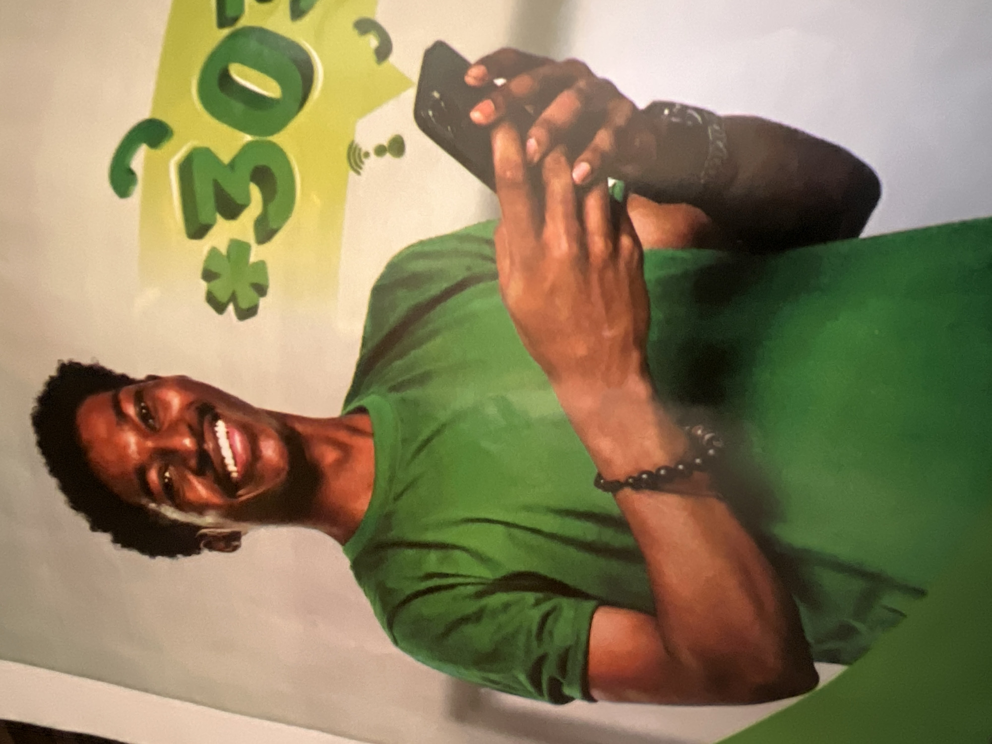 The image is a medium shot of a Black man in a bright green t-shirt, smiling and looking at the viewer. He has short, curly hair and a mustache. He is holding a black smartphone in his right hand, looking down at it. On his left wrist, he wears a bracelet made of dark, round beads. In the background, there is a graphic of a speech bubble with a green asterix and the text "*303#". The overall aesthetic is commercial, likely an advertisement. The scene appears to be indoors, with a plain, light-colored background. There is no visual information to pinpoint the location to Jalingo, Nigeria, or any specific city.