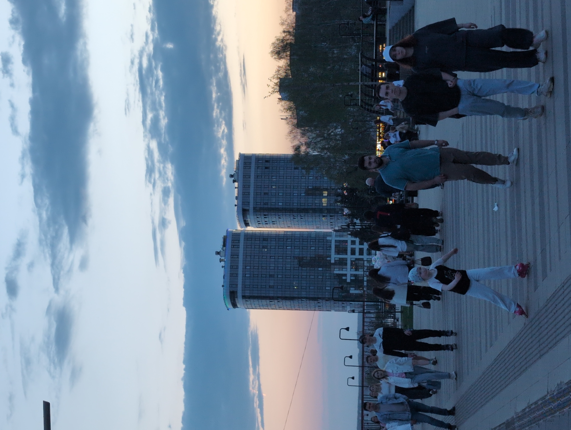 The image shows a gathering of people on a paved area in what appears to be a public space in Izhevsk, Russia. Two high-rise buildings are visible in the background. A group of people are standing on the paved area, some appearing to be posing for a photo. The sky is visible with some clouds. The overall lighting suggests it's either late afternoon or early evening. Some street lamps are present.