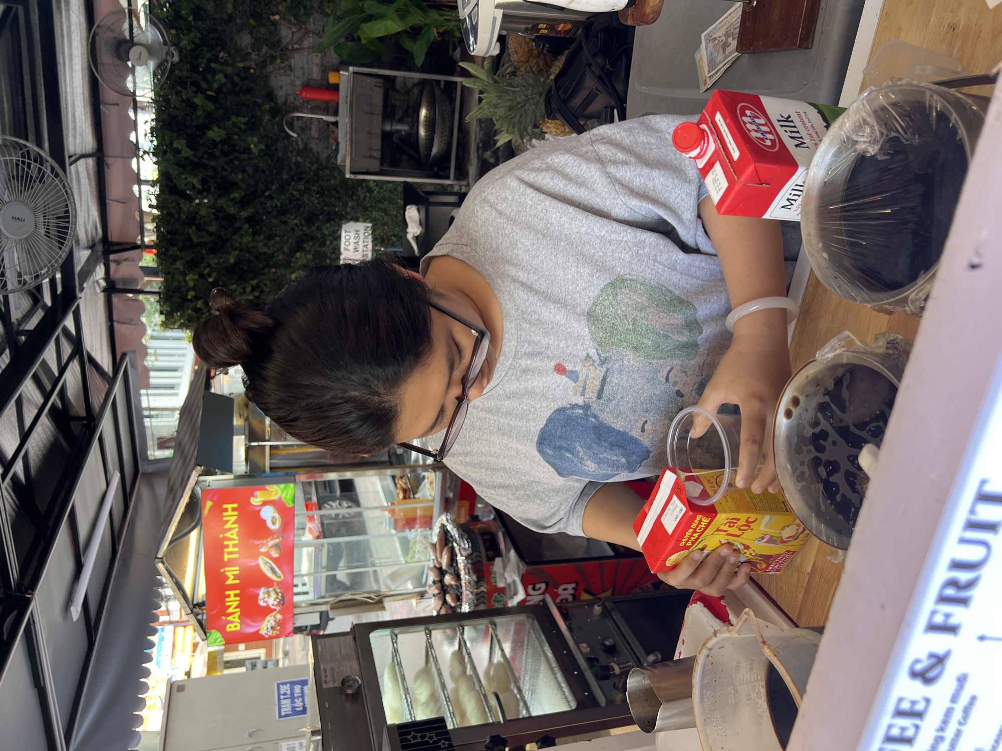 A person, likely a vendor, is shown from a slightly elevated perspective, preparing a drink at a food stall. They are wearing glasses, a grey t-shirt, and have dark hair tied back. The person is in the process of pouring liquid from a yellow carton, labeled "Tai Loc" and "CHUYÊN DÙNG PHA CHẾ" (Specialized for Preparation), into a clear plastic cup. On the wooden counter in front of them, a large pot with a clear lid contains a dark liquid, possibly a coffee or tea base, and a red and white carton of "Milk" (marked "Long Life, Full Fat") is also present. A white, translucent bracelet is visible on the person's left wrist.
The setting is an outdoor or semi-outdoor market stall, indicated by fans, lush green foliage in the background, and various signs. A prominent red sign advertises "BÁNH MÌ THÀNH" (Banh Mi Thanh), suggesting a Vietnamese eatery. Another blue sign reads "TRAM T.29E LỘC THỌ". To the right, a vertical sign partially displays "COFFEE & FRUIT", with "kem muối" (salted cream) and "coffee" also visible. An unexpected sign on a wall near the foliage points to a "FOOT WASH STATION". Bright lighting indicates it is daytime. In the lower left, a display case with what appears to be baked goods or snacks is visible. The scene captures a moment of food preparation in a bustling street food environment, consistent with the provided location context of Nha Trang Ward, Vietnam.