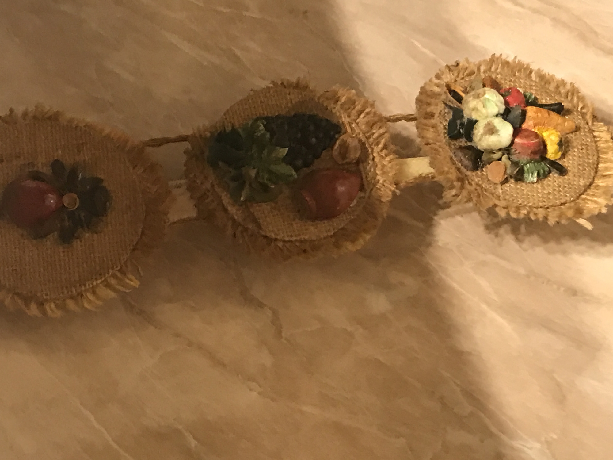 The image shows a garland-like decoration consisting of three burlap circles, each adorned with miniature fruits and vegetables. The left circle features a red fruit with dark leaves, the center circle showcases grapes, an apple, and green leaves, and the right circle displays garlic, a carrot, a red apple, and other vegetables. The circles are connected by a thin rope. The background is a textured surface in shades of beige and brown.
There is no information indicating that this image was taken in Yerevan, Armenia, or that any specific people are involved.