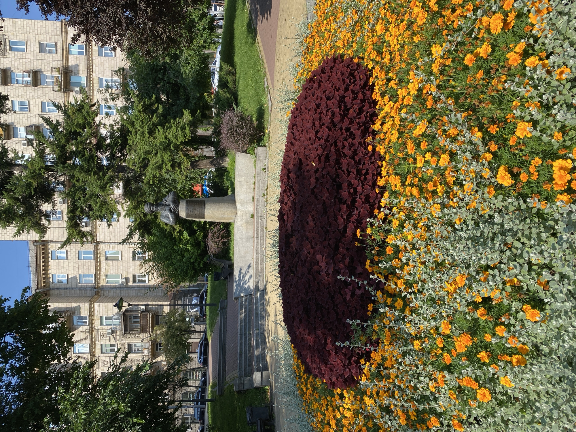 An eye-level, full shot captures a public park area with a large flowerbed in the foreground and a monument and building in the background. The flowerbed is circular, with a dark red, bushy plant in the center surrounded by bright orange flowers and silvery-green foliage. The flowerbed is bordered by a yellow paved walkway. Behind the flowerbed, a gray stone monument with a bust of a man wearing a turban stands on a pedestal, positioned on a raised concrete platform. Steps lead up to the platform from a paved pathway. Beyond the monument, a lush green lawn is visible, with trees and a tall, light-colored building with multiple windows and balconies in the distance. The sky is clear and blue. The scene appears to be well-maintained and bathed in sunlight. There are no people visible in the immediate foreground.